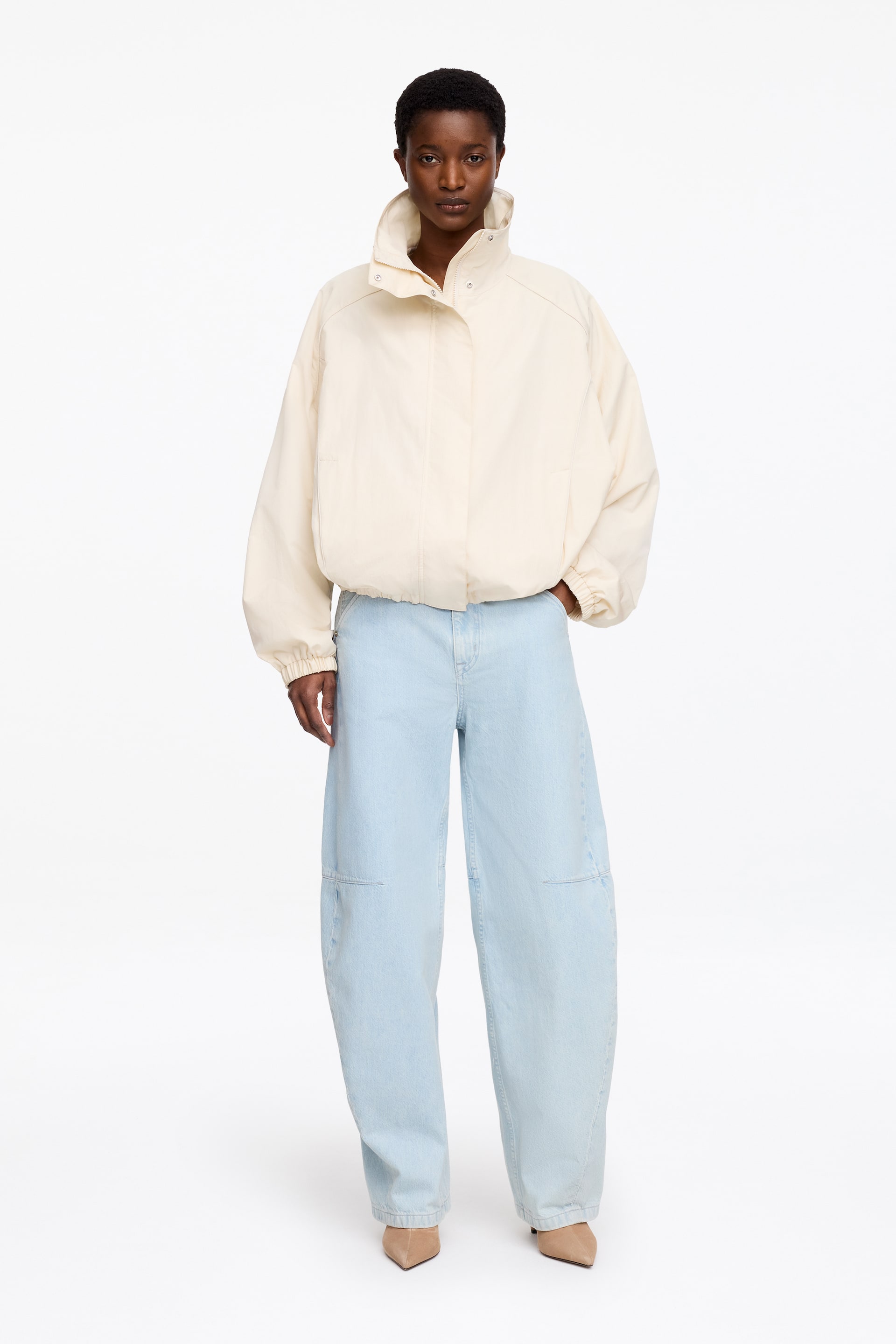 Visa större bild: En krämfärgad bomberjacka med hög ståkrage och tryckknappdetaljer samt dold frontstängning. Den har voluminösa långa ärmar med rynkade muddar och en elastisk fåll, vilket ger en avslappnad passform framifrån.