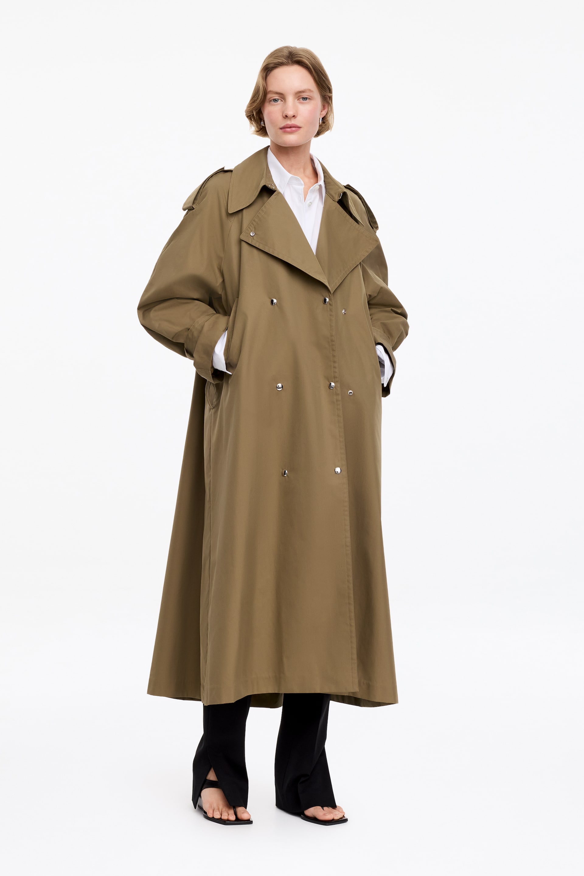 Grotere afbeelding bekijken: Een olijfgroene double-breasted trenchcoat wordt gedragen door het model, die met haar handen in de zijzakken staat. De jas is ontworpen met brede revers, epauletten, stormflappen en zilveren knopen, en is gestyled over een witte blouse, zwarte broek en zwarte sandalen.