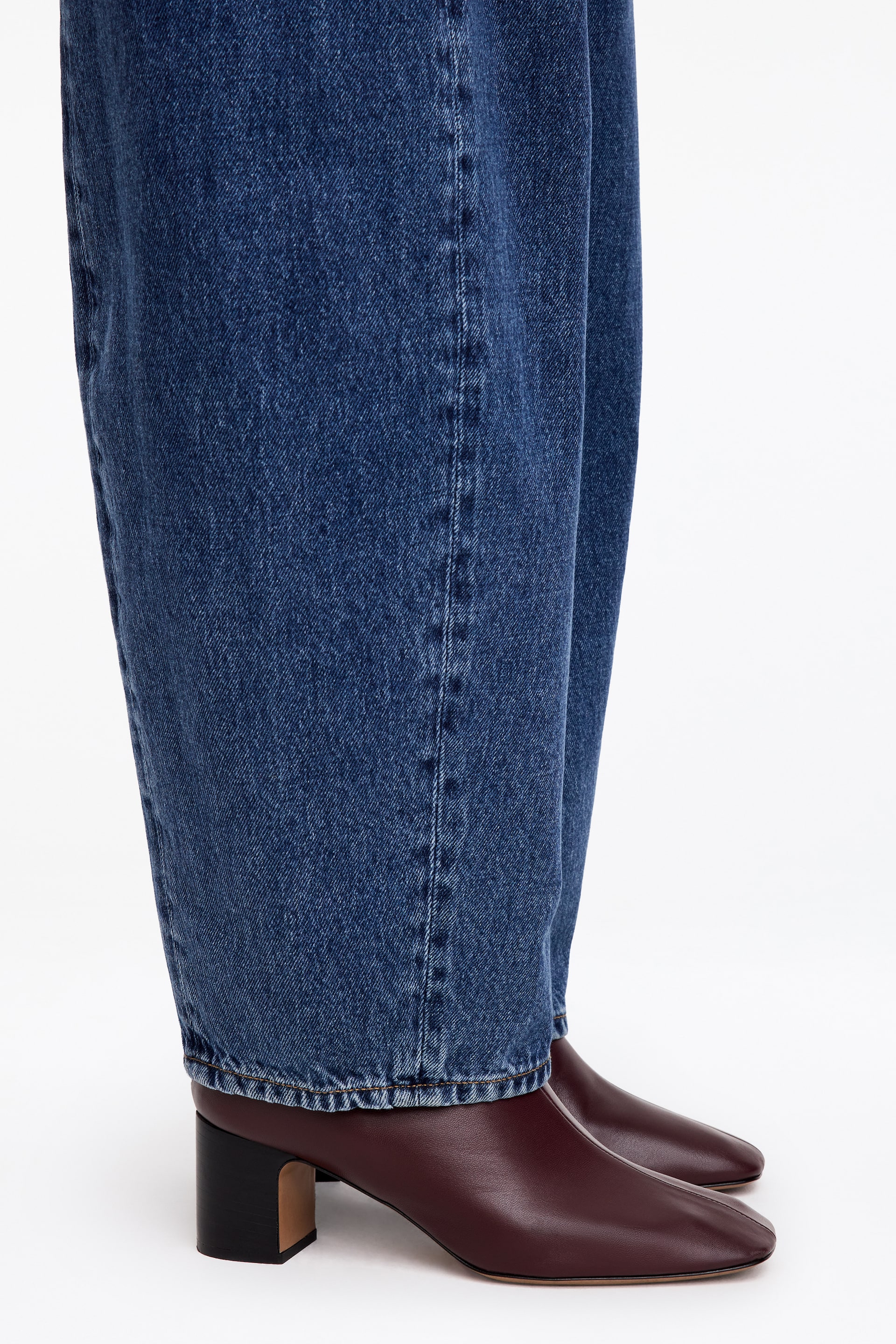 Visa större bild: Mörkt vinröda ankelstövlar i läder med fyrkantig tå och en grov svart klack bärs av modellen under vida blå jeans i denim.