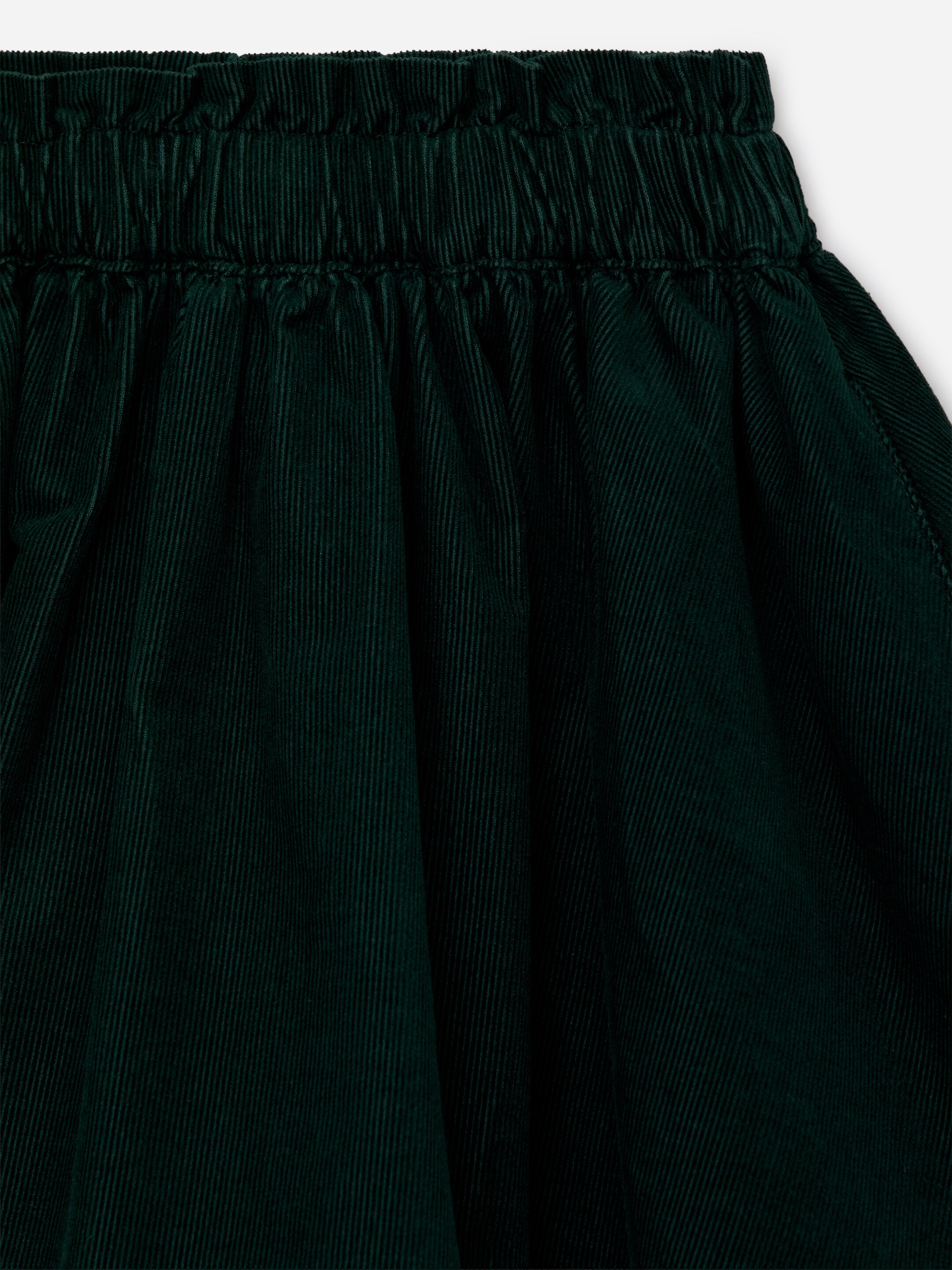 View larger image: A dark green corduroy skirt is displaying an elasticated paperbag waist. The corduroy fabric is showing vertical wales and soft gathers below the waistline.