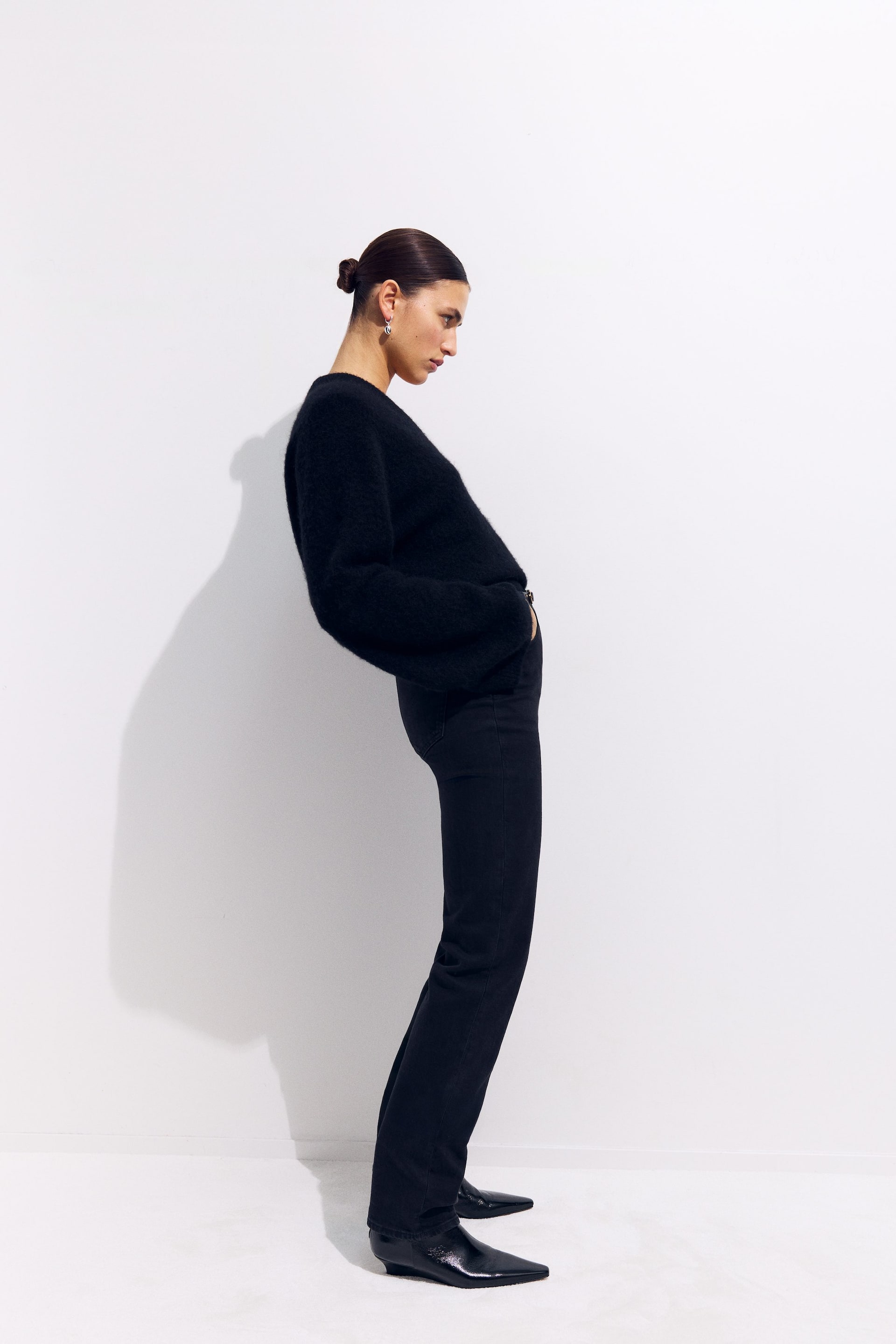 Visa större bild: En svart crewneck-tröja med en luddig textur och avslappnad passform visas från sidan. Den har långa ärmar och är stylad med svarta raka byxor som täcker svarta spetsiga stövlar.