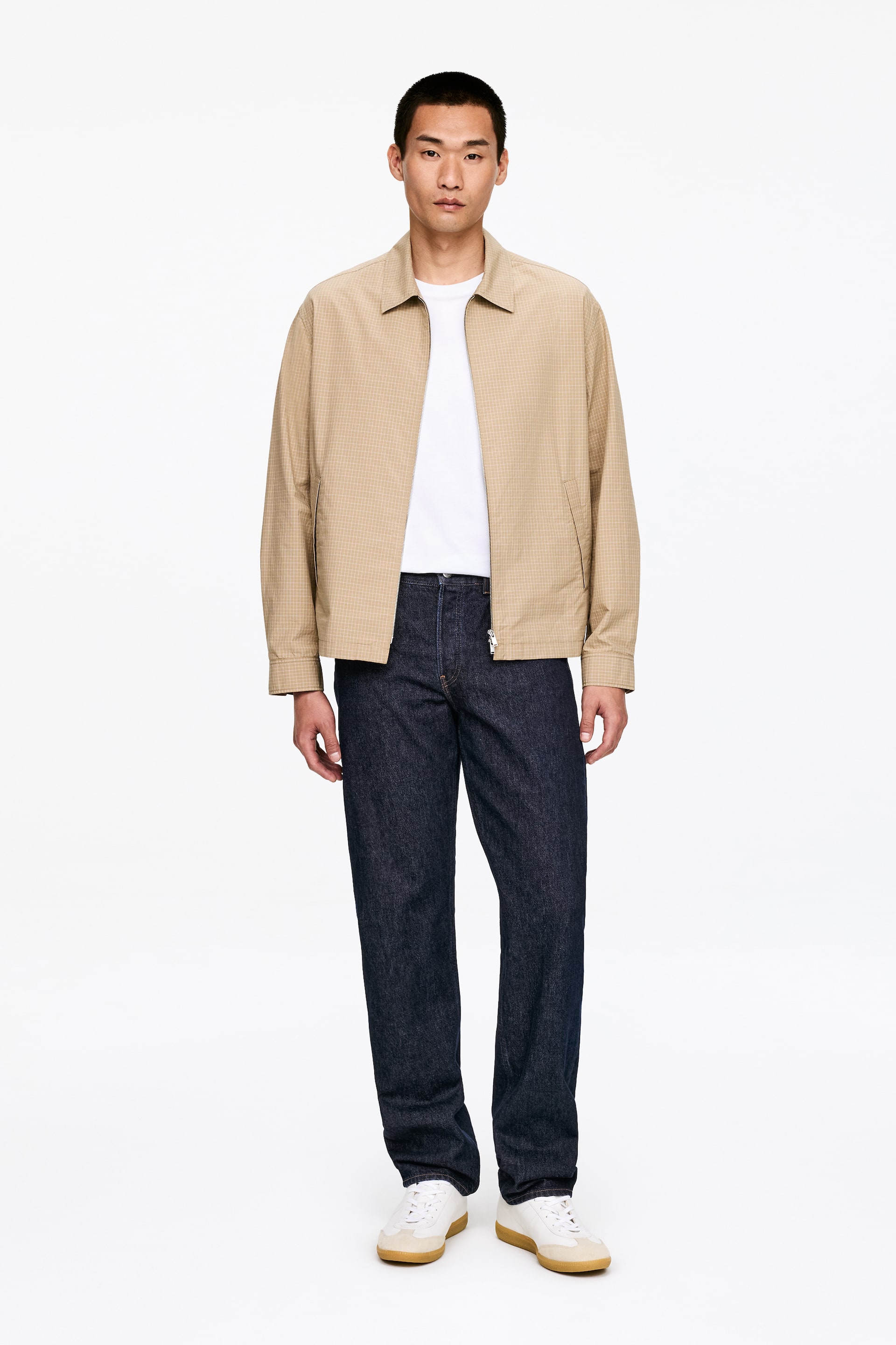 Ver imagen más grande: Una chaqueta beige presenta un sutil patrón de microcuadros, un cuello de punta y un cierre frontal con cremallera. Se lleva abierta sobre una camiseta blanca, revelando bolsillos laterales y mostrando su ajuste relajado.