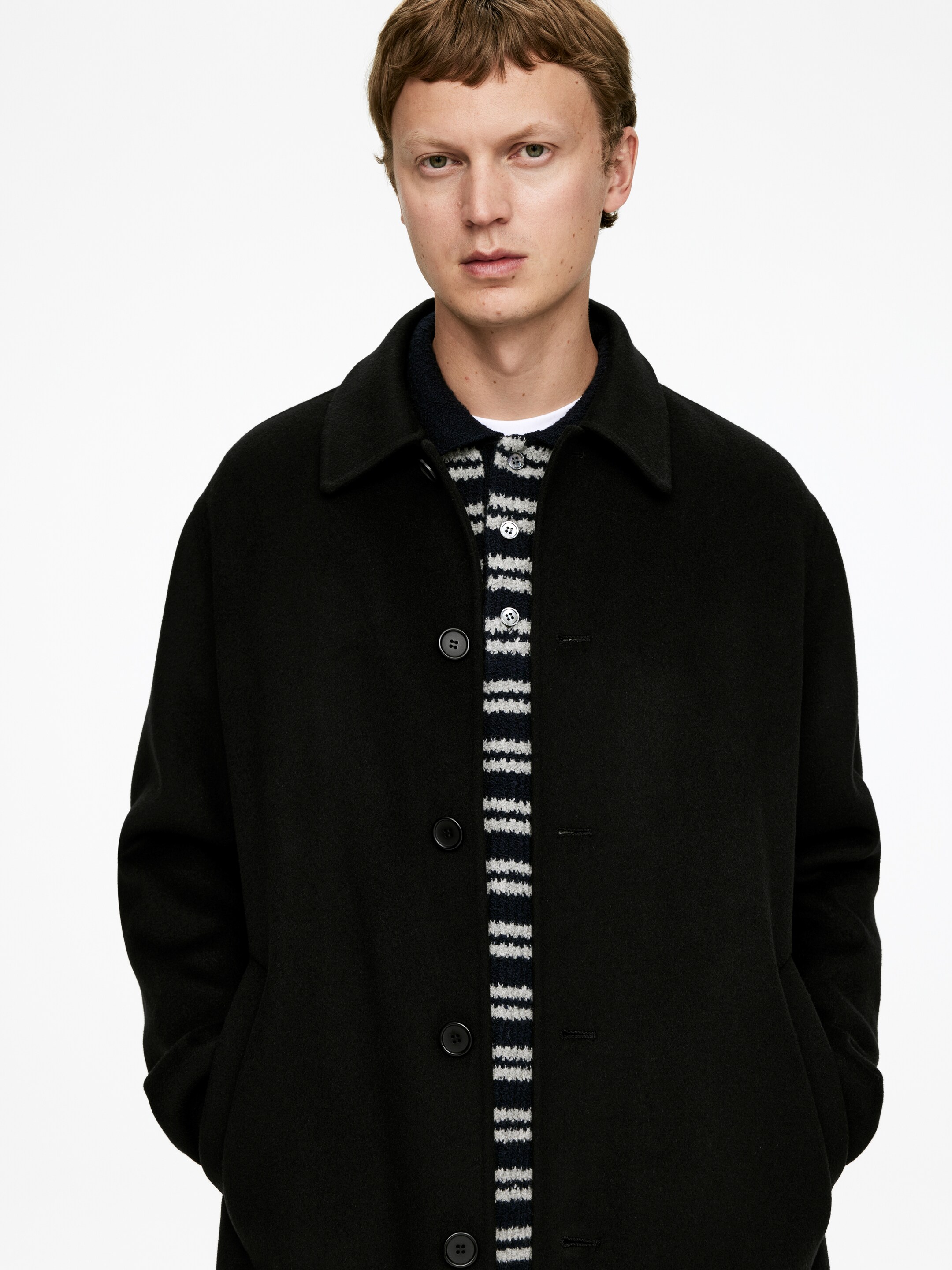 Ingrandisci l'immagine: Un cappotto nero è presentato frontalmente, caratterizzato da un classico colletto a punta e tre bottoni frontali visibili. Un maglione a righe è stratificato sotto il cappotto aperto e le mani di chi lo indossa sono appoggiate nelle tasche laterali del cappotto, indicando una vestibilità rilassata.