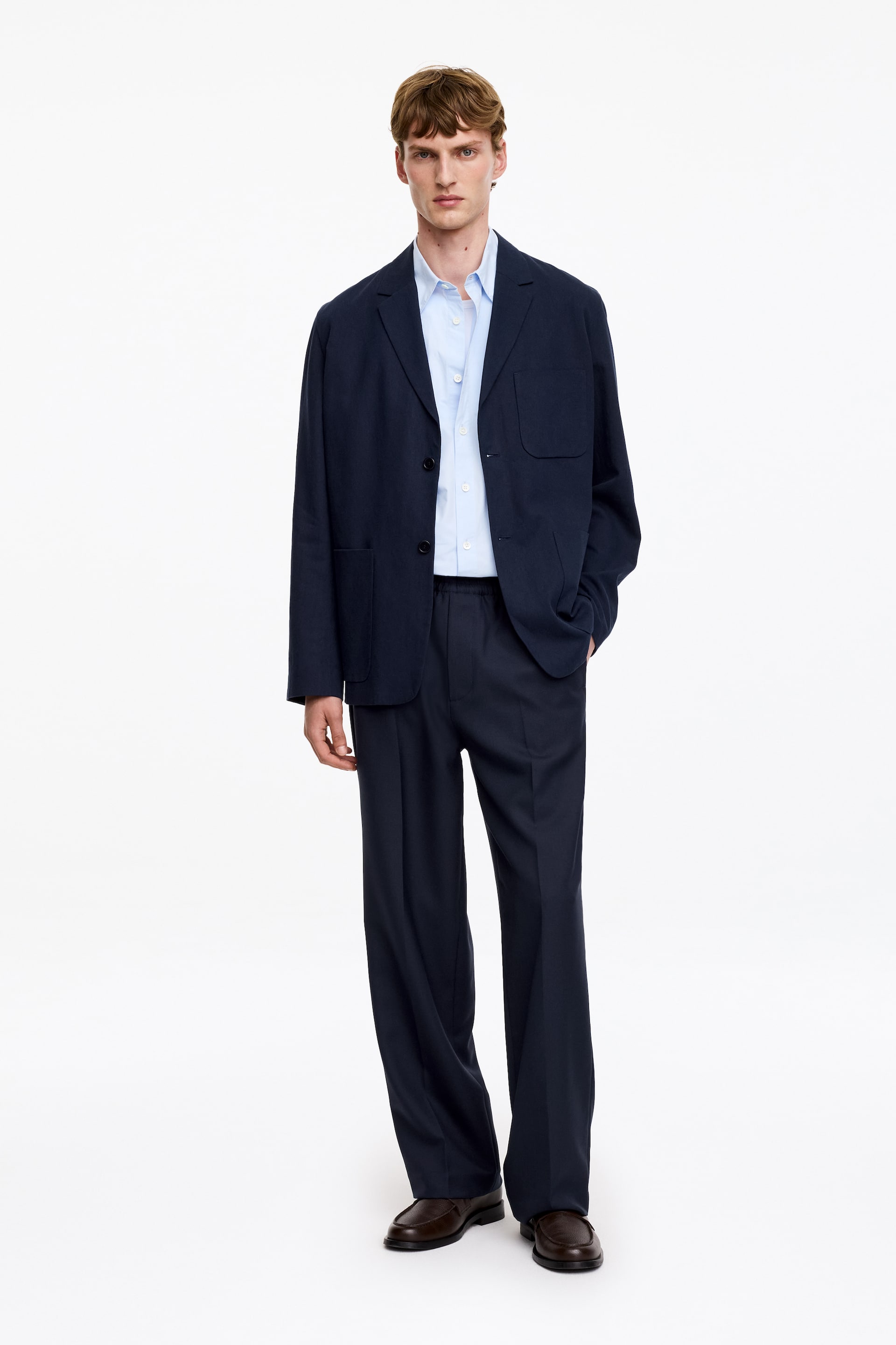 Ver imagem maior: Um blazer azul-marinho de corte descontraído exibe lapelas de entalhe e dois bolsos frontais de chapa, com um dos seus dois botões frontais abotoado, usado sobre uma camisa azul-clara de botões. O blazer complementa calças largas azul-marinho que caem sobre mocassins castanho-escuros.