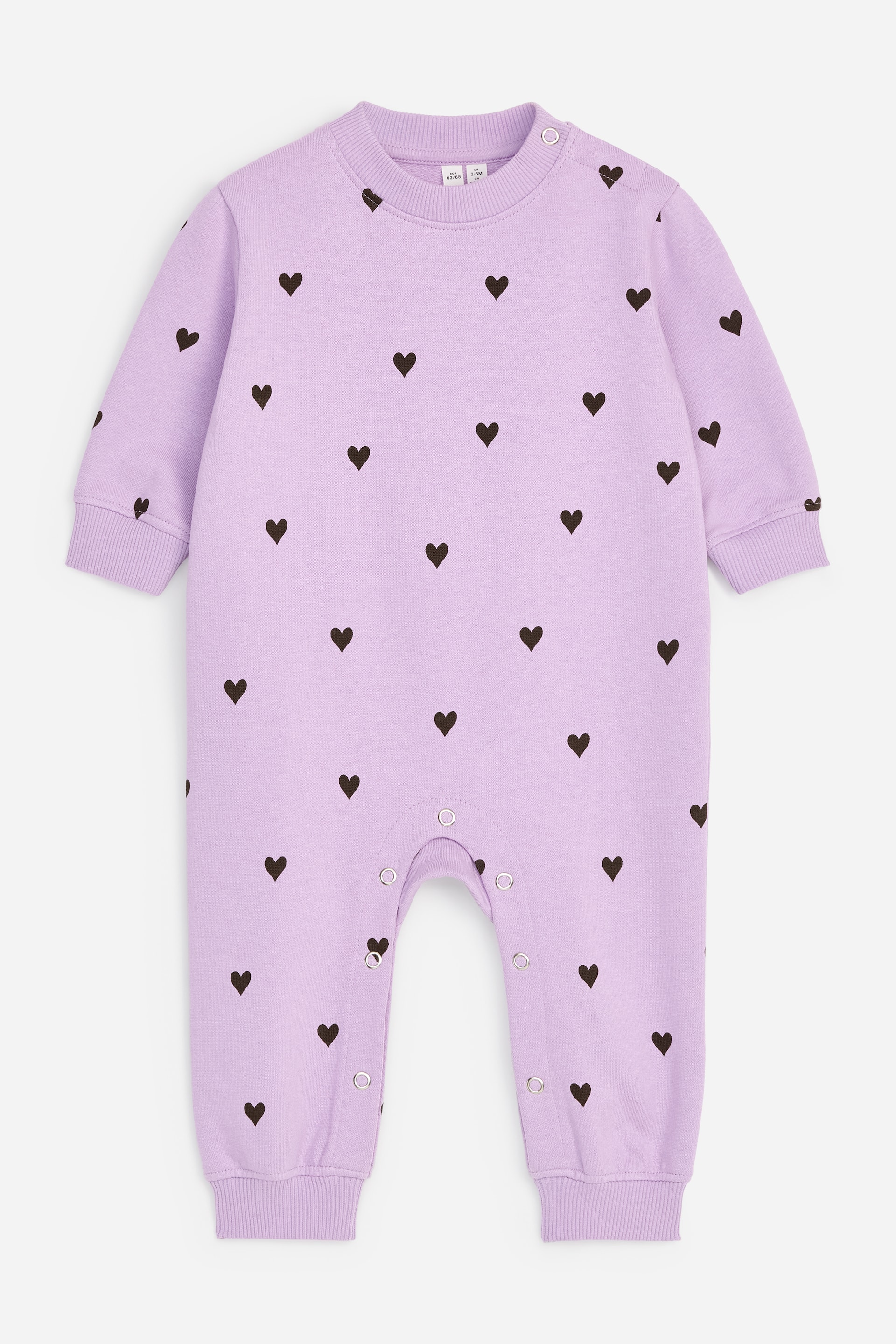 View larger image: A light purple baby romper is laid flat, showcasing a scattered pattern of small, dark brown hearts. It is designed with long sleeves and legs, both finished with ribbed cuffs, and fastens with snap buttons along the left shoulder and inner legs.