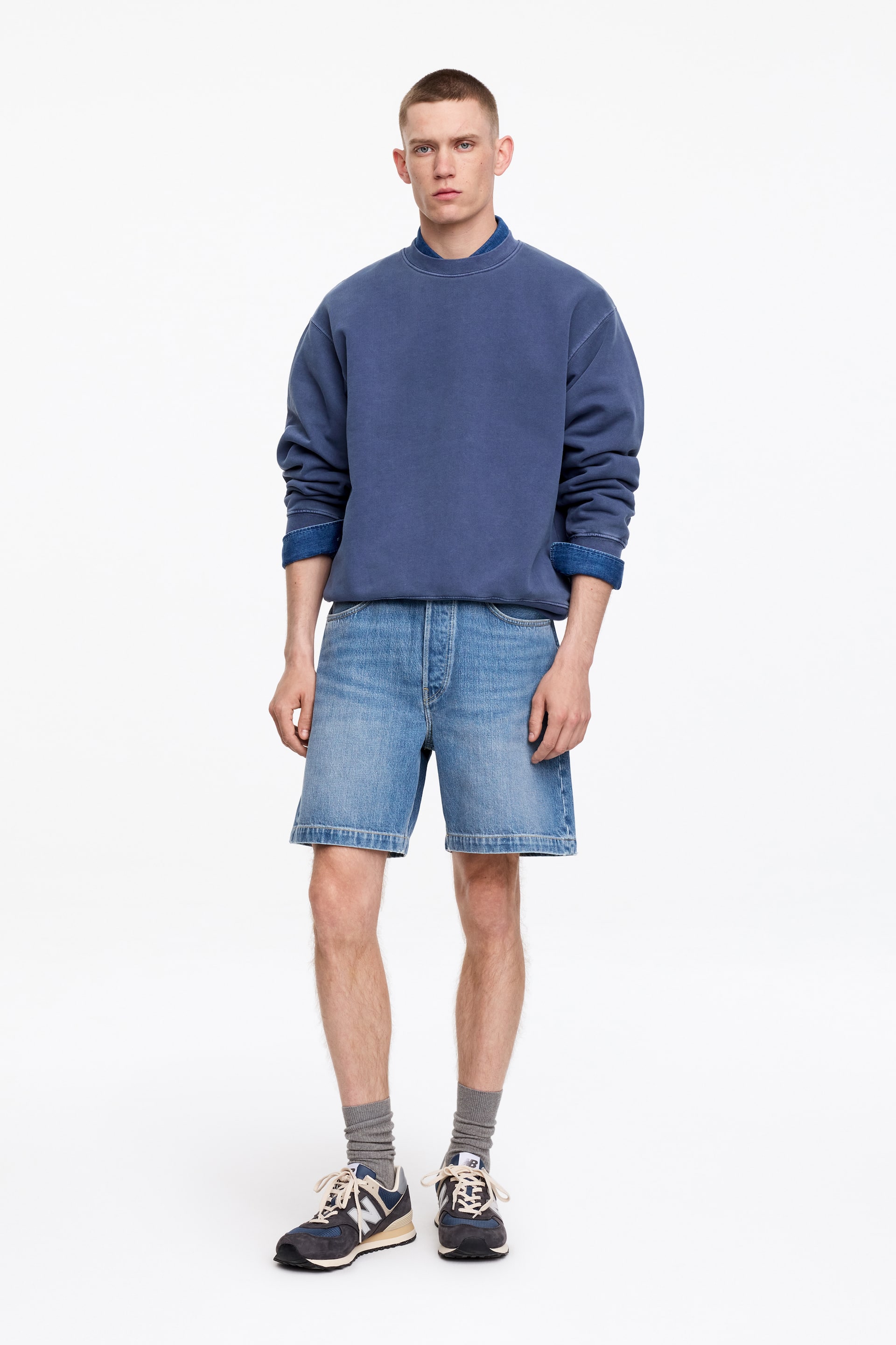 View larger image: A faded blue crewneck sweatshirt is displayed from the front, featuring a ribbed collar, ribbed cuffs, and a ribbed hem. A blue collared shirt is visible layered underneath, its collar showing above the neckline and its cuffs extending beyond the sweatshirt's cuffs.