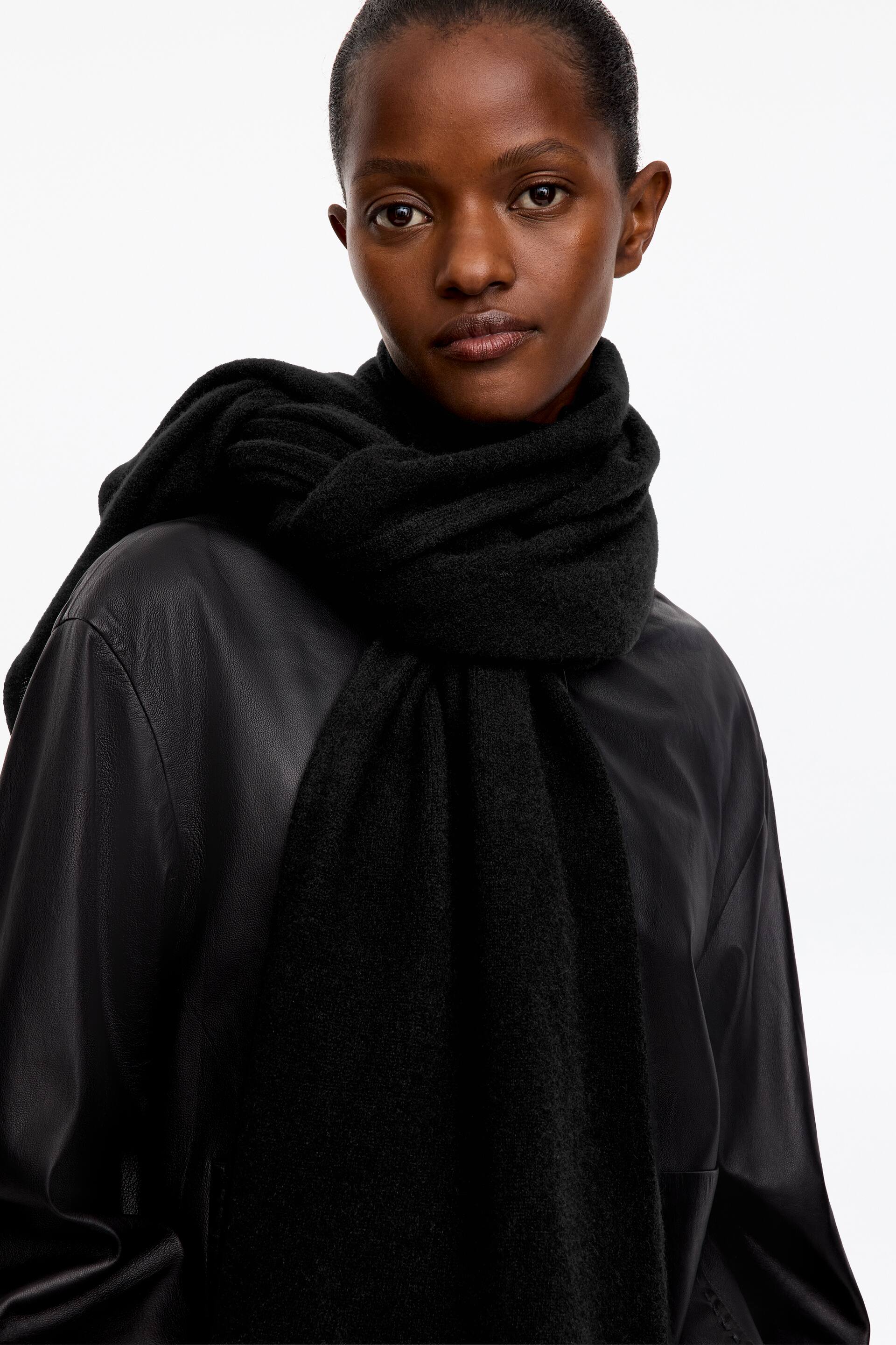View larger image: A black knitted scarf with a soft, brushed texture is being worn by the model, wrapped around the neck with one end hanging down at the front and the other draped over the shoulder. The accessory is layered over a black leather jacket.