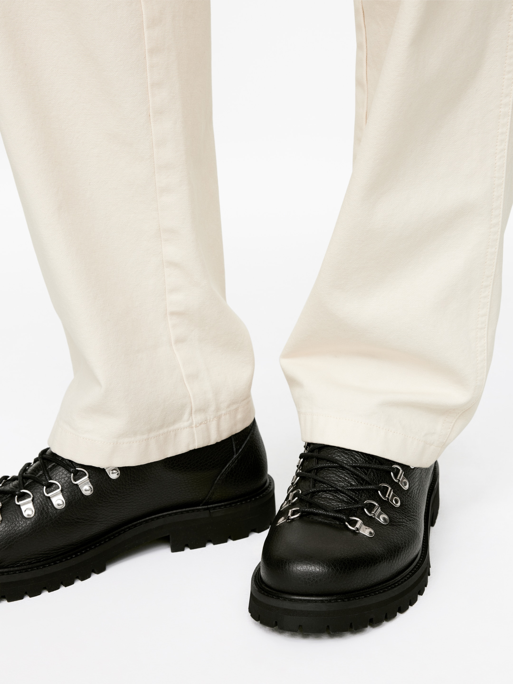 Grösseres Bild ansehen: Schwarz genarbte Lederspaziergenschuhe werden vom Model getragen, das sie mit hellen Hosen kombiniert. Die Schuhe zeigen ein Schnürdesign mit schwarzen gewachsten Baumwollschnürsenkeln und silberfarbenen Metallschlaufenösen und stehen auf einer dicken profilierten Gummisohle.