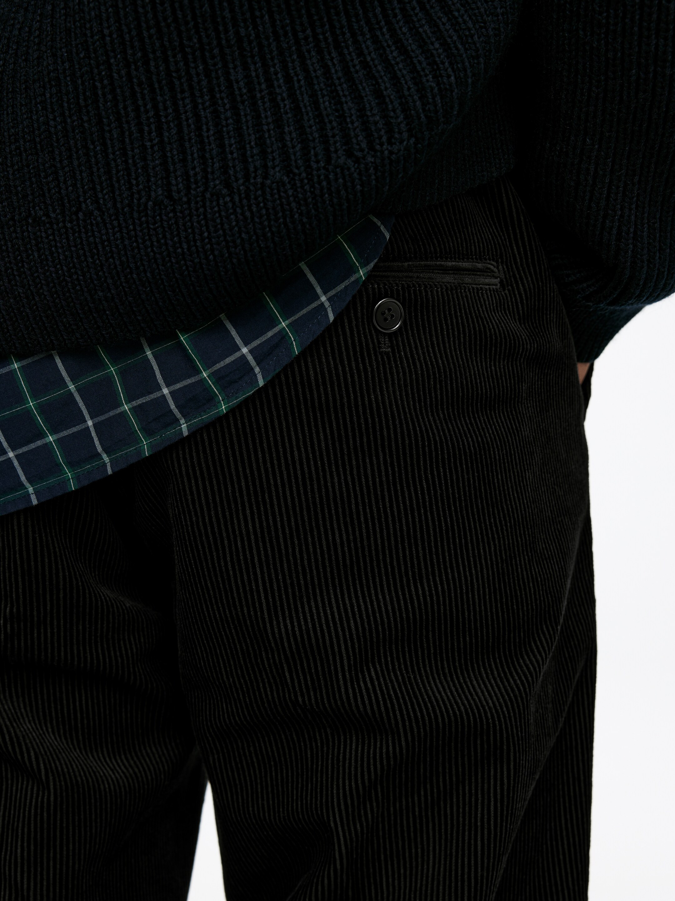 Größeres Bild anzeigen: Schwarze Cordhosen werden vom Model getragen. Eine paspelierte Gesäßtasche mit Knopf ist an den Hosen sichtbar. Ein dunkelblauer gerippter Pullover wird über einem karierten Hemd getragen, und die Hand des Models ruht in der Nähe der Gesäßtasche.
