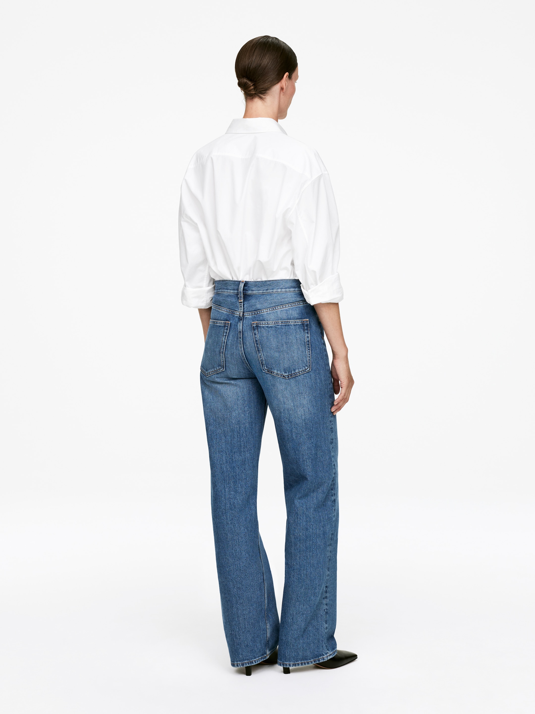 Ingrandisci l'immagine: I jeans in denim blu presentano una vestibilità a gamba larga, visti da dietro, con due tasche posteriori e un leggero scolorimento sulle cosce. Una camicia bianca è infilata nella loro vita alta.
