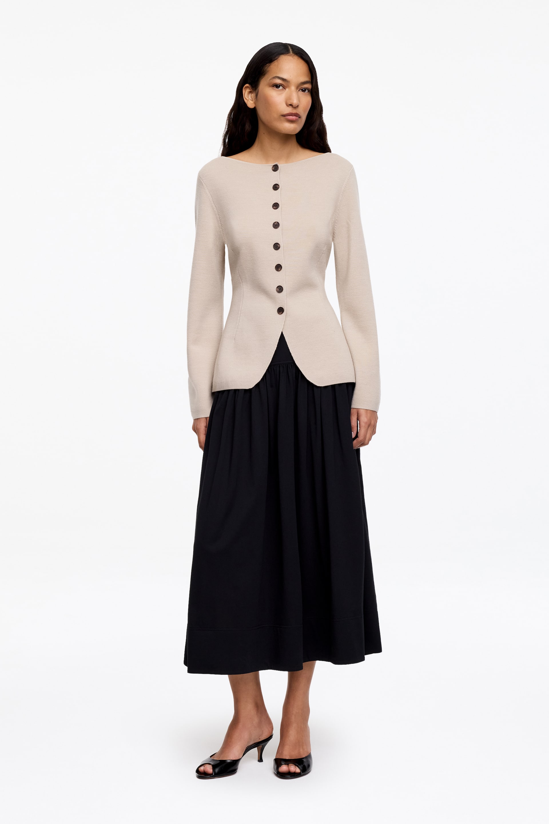 Grotere afbeelding bekijken: Een licht beige aansluitende gebreide top met een boothals en lange mouwen, met zeven donkere knopen langs de voorkant. De top heeft een aansluitende taille, een licht uitlopende, gebogen zoomlijn en wordt gedragen over een zwarte rok.