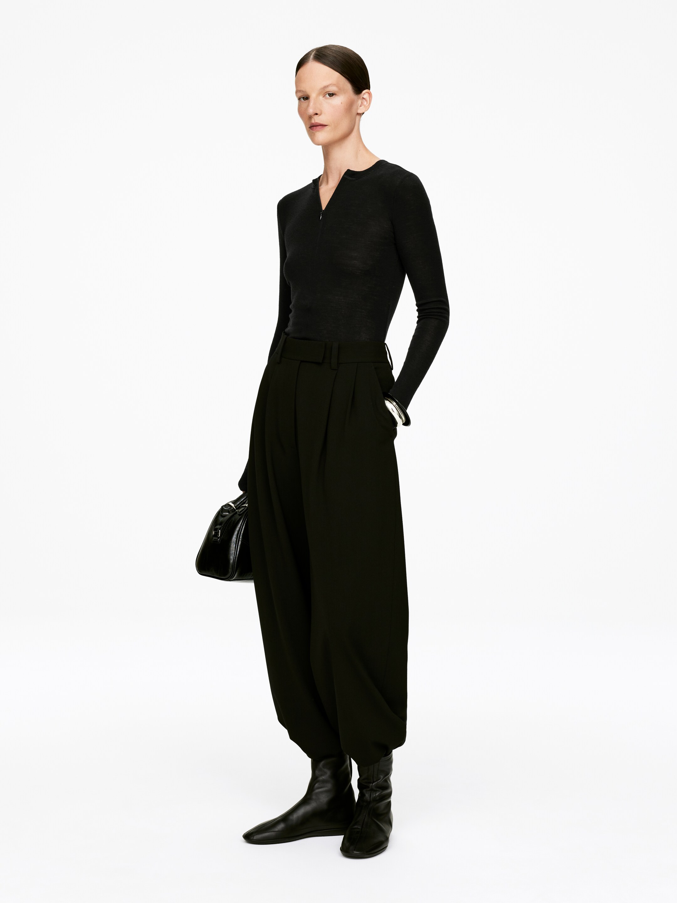 Agrandir l'image: Un haut noir à manches longues avec une fermeture éclair partielle à l'encolure est associé à un pantalon noir taille haute, qui présente un design ample à jambes larges avec des plis à la taille et se rétrécissant aux chevilles. Des bottines noires plates à bout pointu complètent l'ensemble, avec un sac à main structuré noir tenu à la main.