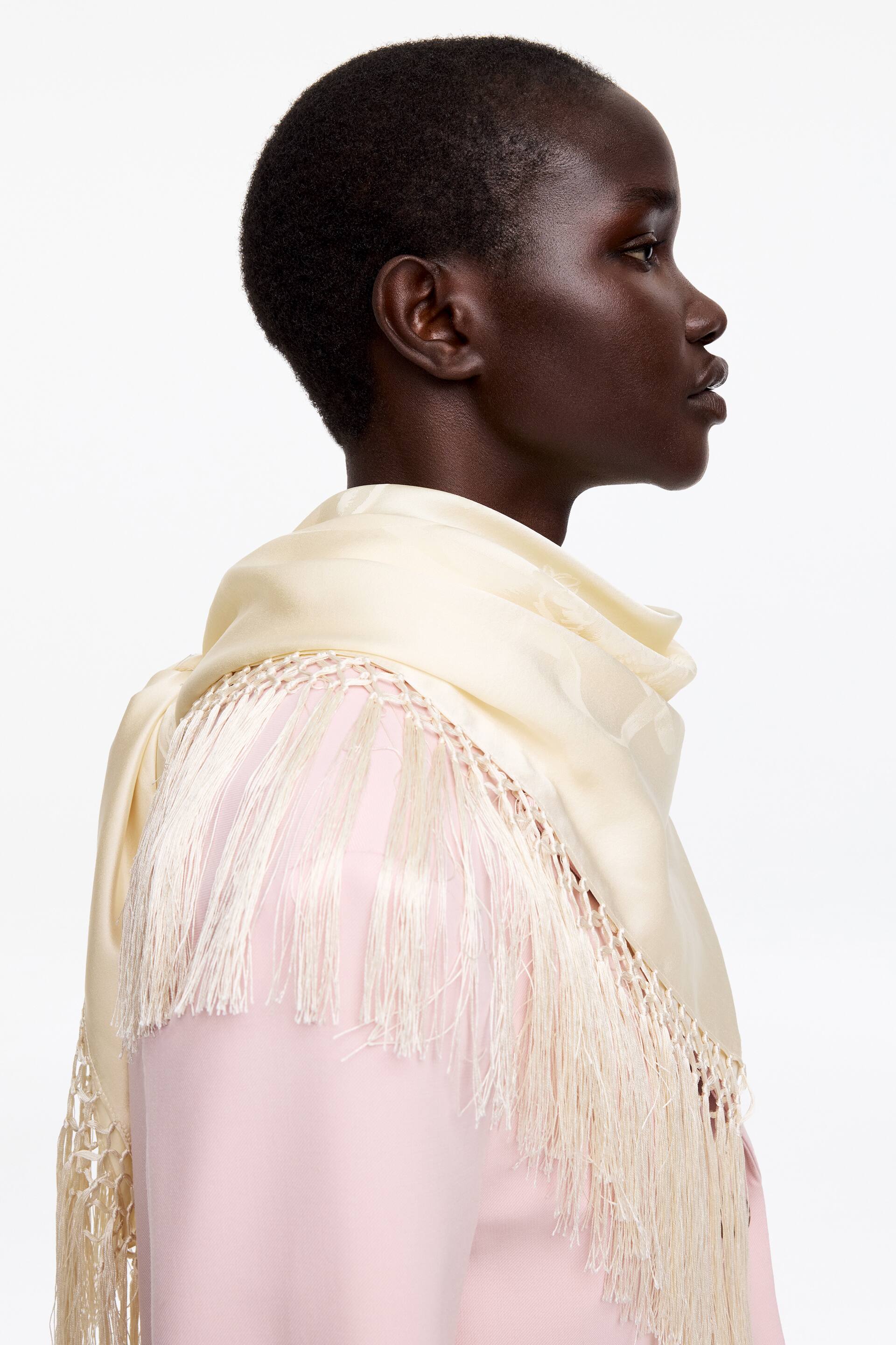 View larger image: Silk Scarf. A cream silk scarf, showcasing a subtle tone-on-tone pattern and lengthy fringe, is elegantly draped over the model's shoulders, complementing a light pink garment underneath.