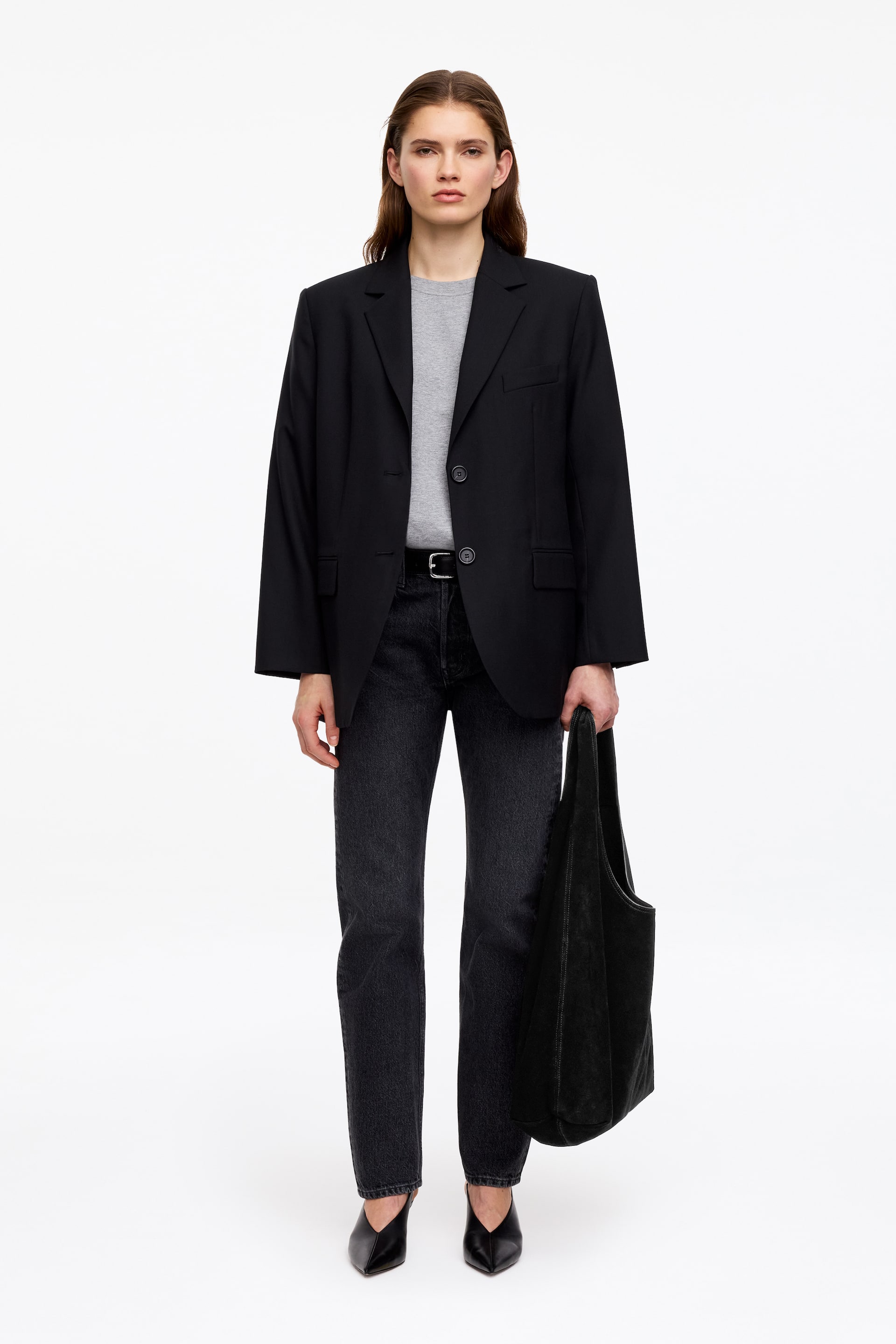 Ver imagem maior: Um blazer preto é apresentado de frente, com lapelas em bico e fecho de dois botões num estilo single-breasted, com um botão abotoado. Apresenta ombros estruturados, um corte descontraído, dois bolsos frontais com aba e um bolso no peito, sobreposto a uma t-shirt cinza-clara de gola redonda.