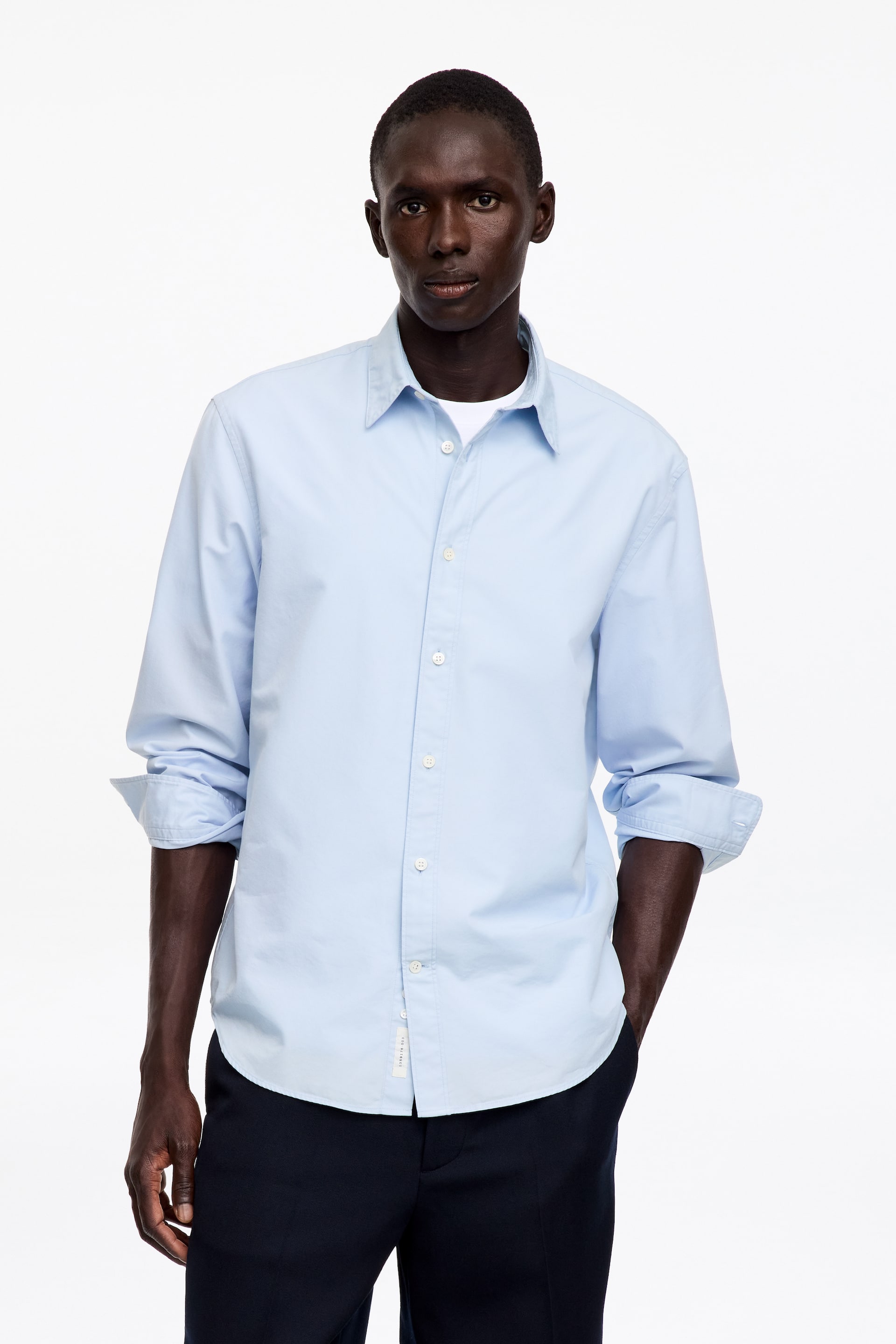 Ver imagen más grande: La modelo lleva una camisa azul claro de manga larga con cierre de botones en la parte delantera. Presenta un cuello clásico y botones blancos, y las mangas están remangadas. La camisa se lleva sobre una camiseta interior blanca.