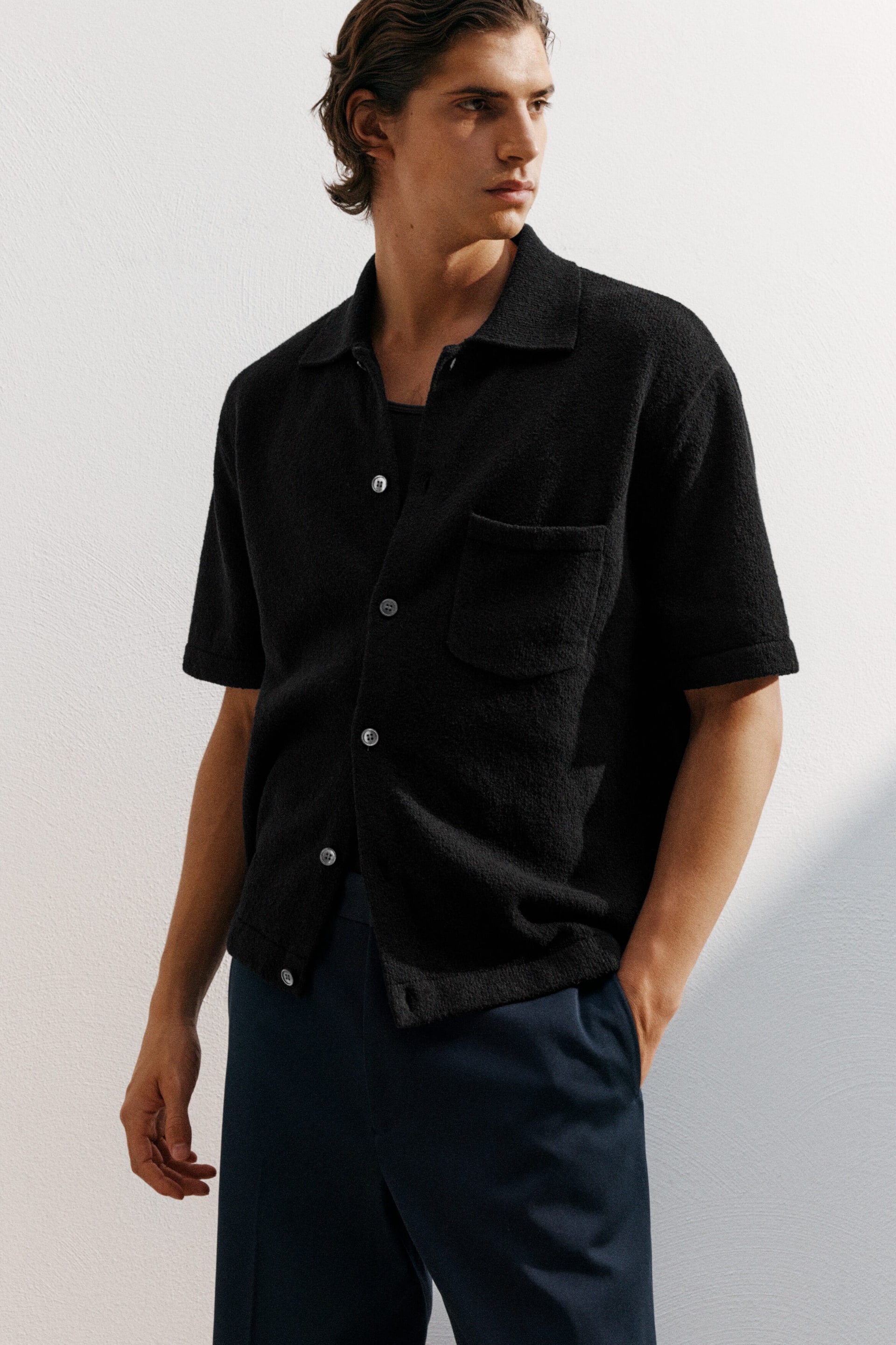 Ver imagen más grande: Una camisa de punto texturizado de manga corta en color negro presenta un cuello tipo campamento, cierre frontal con botones de color claro y un único bolsillo de parche en el pecho izquierdo. Se lleva abierta sobre una camiseta interior más oscura, mostrando su ajuste relajado desde un ángulo ligero.