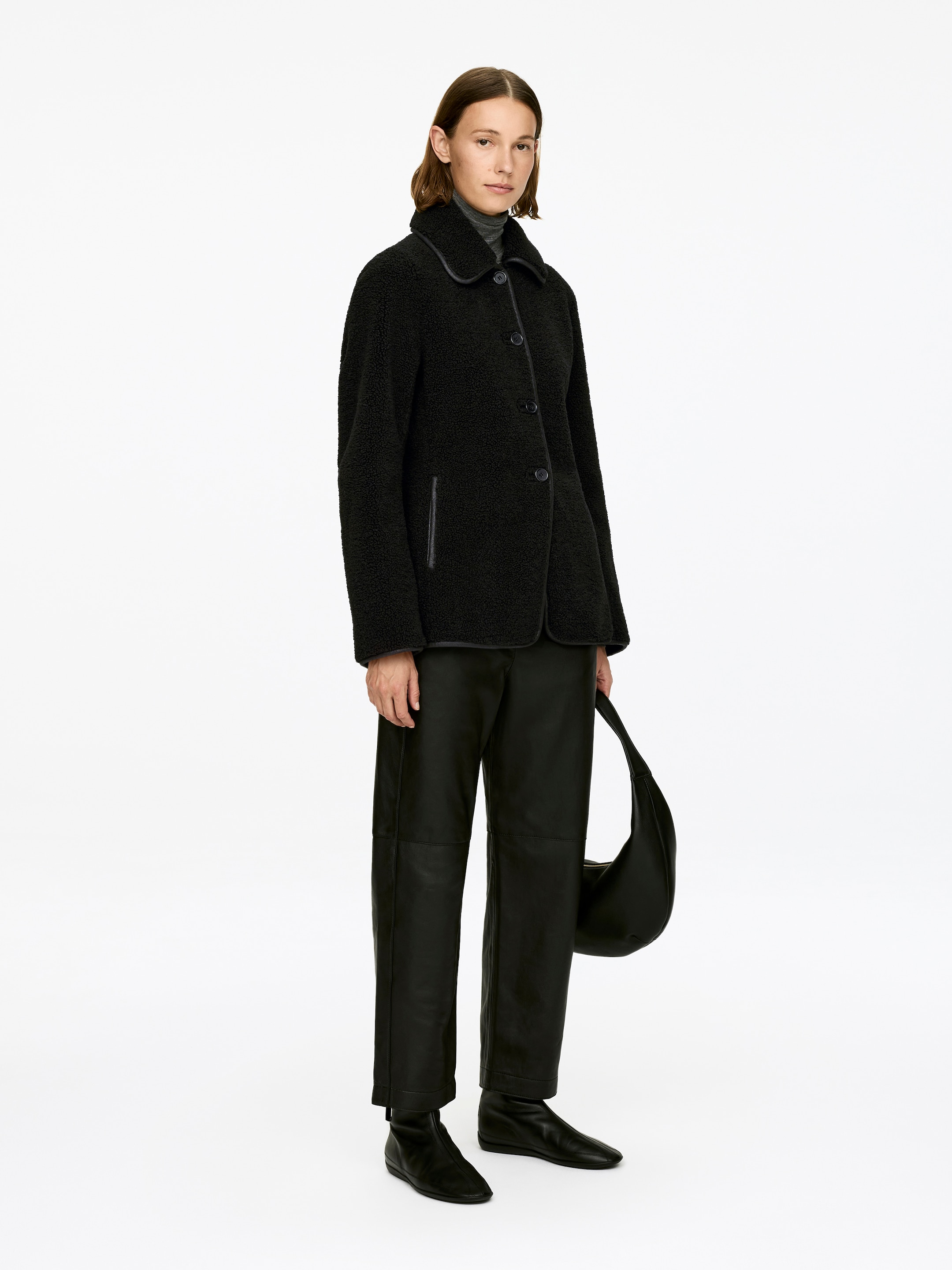 View larger image: A black single-breasted pile jacket, featuring a classic collar, single welt pockets, and contrasting piping along its edges, is being worn by the model. It is layered over a grey turtleneck.