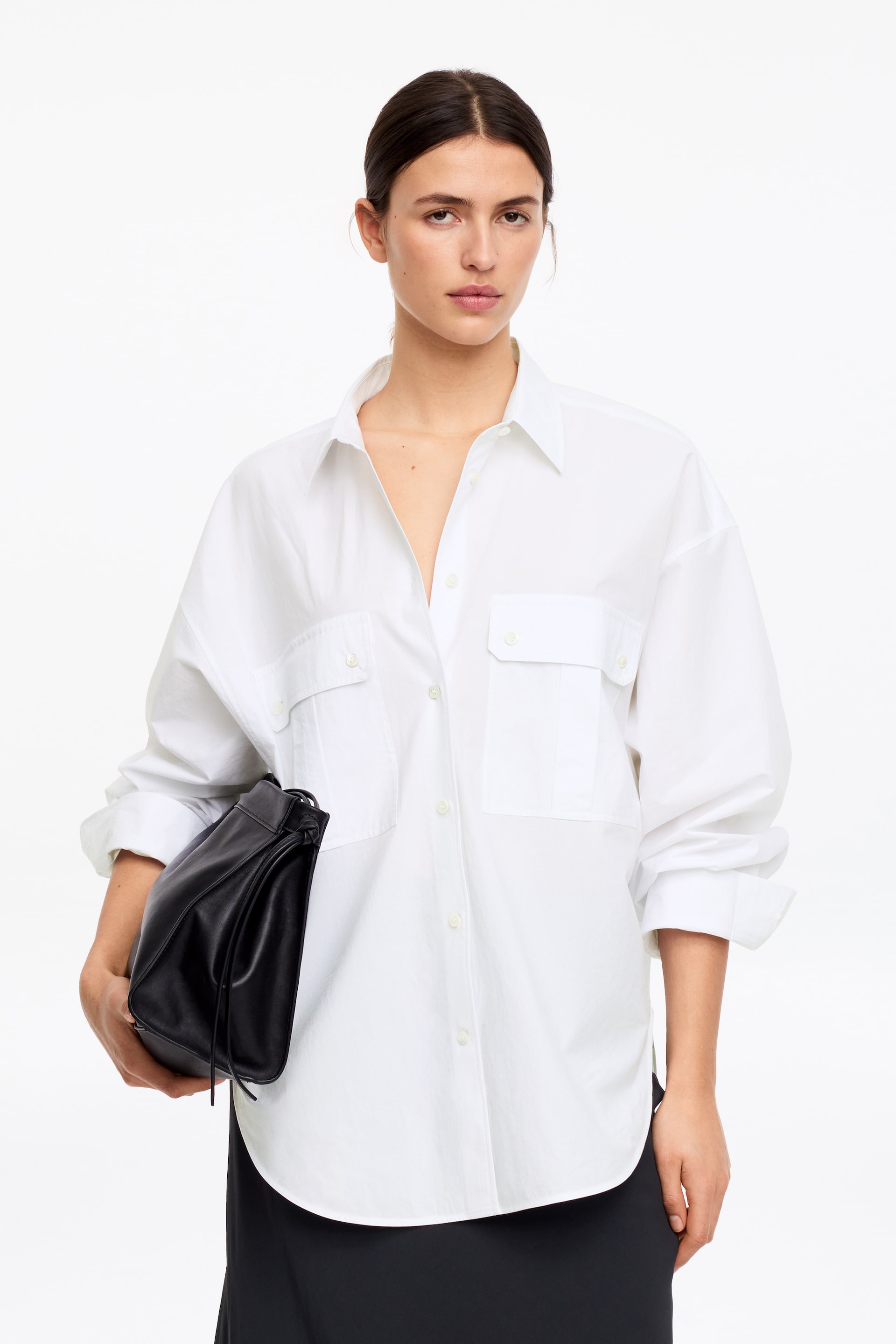 Nagyobb kép megtekintése: Egy fehér, túlméretezett ing klasszikus hegyes gallérral, látható elülső gombos záródással és két nagy mellzsebbel rendelkezik. Hosszú ujjai a mandzsettánál fel vannak hajtva, és lekerekített szegélye is látható.