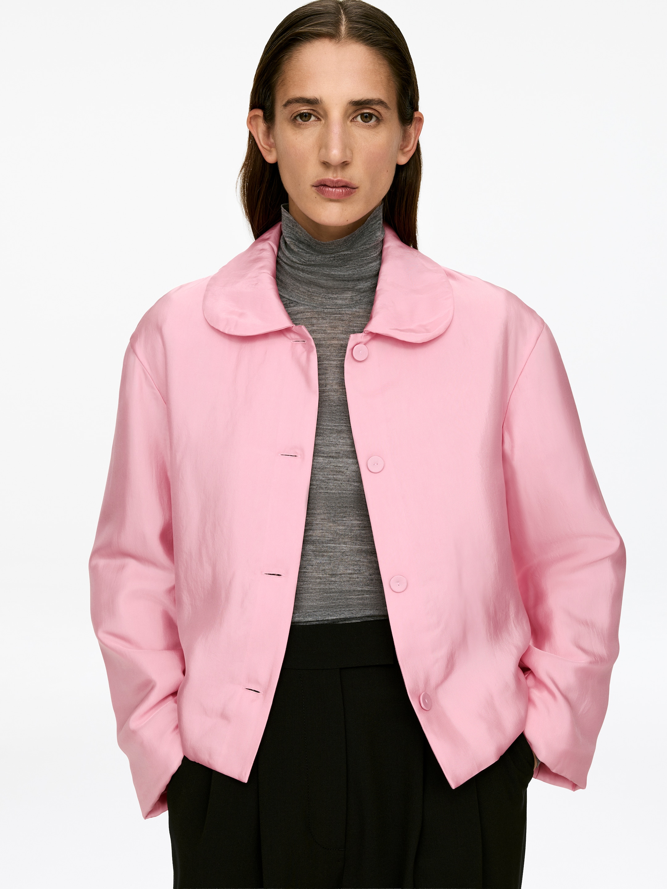 Vis større bilde: En rosa polstret overskjorte med avrundet krage og stofftrukne knapper, med en vanlig passform, bæres åpen av modellen. Den er lagd over en grå turtleneck, og modellens hender hviler i bukselommene, med overskjorten drapert over dem.