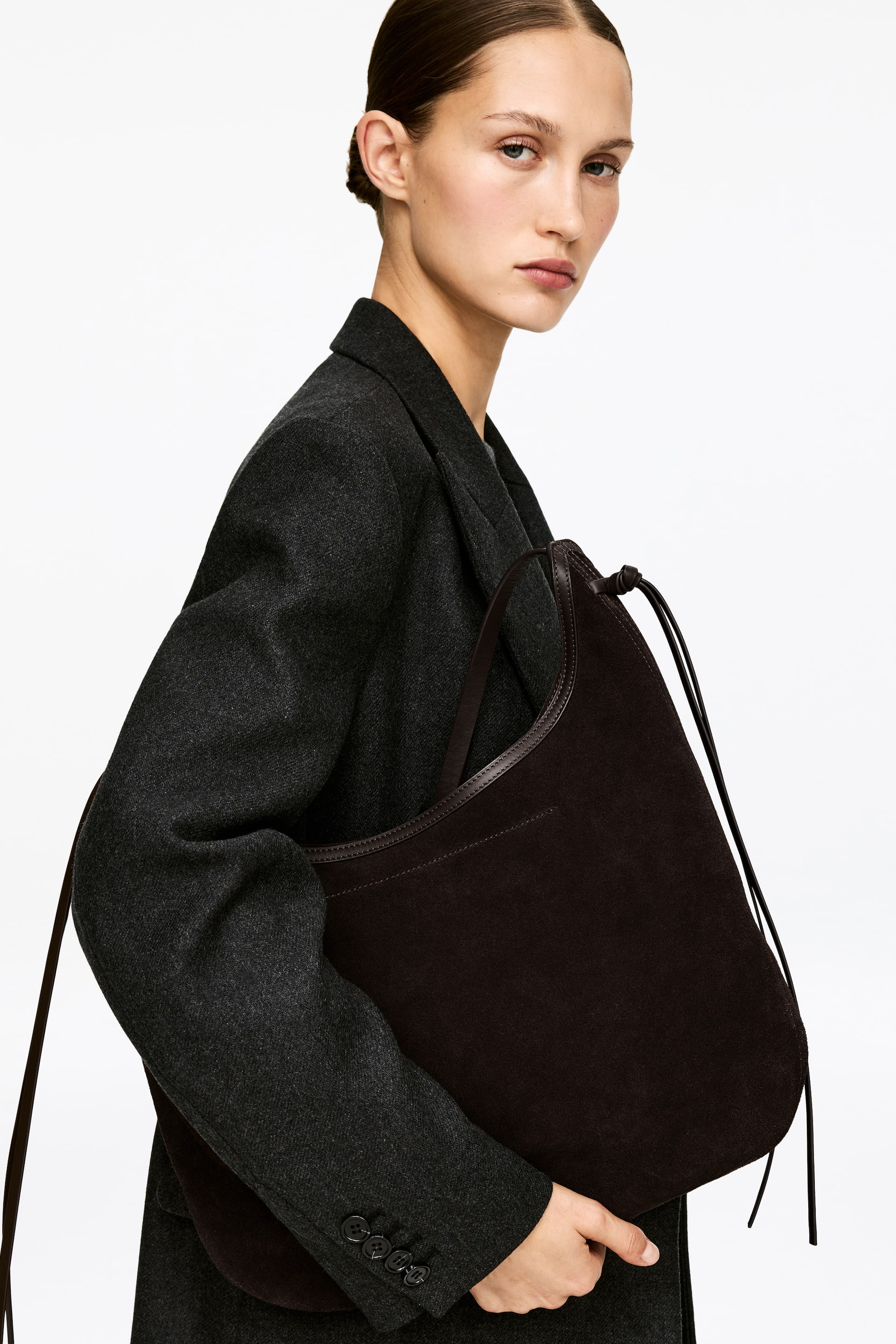 View larger image: A dark brown hobo bag, crafted from a soft, textured material reminiscent of suede, is showcased from a slightly angled side perspective. It features a slender, dark leather shoulder strap, a coordinating leather trim along its top edge, and thin cord-like ties visible on its side, while resting against a dark grey blazer.