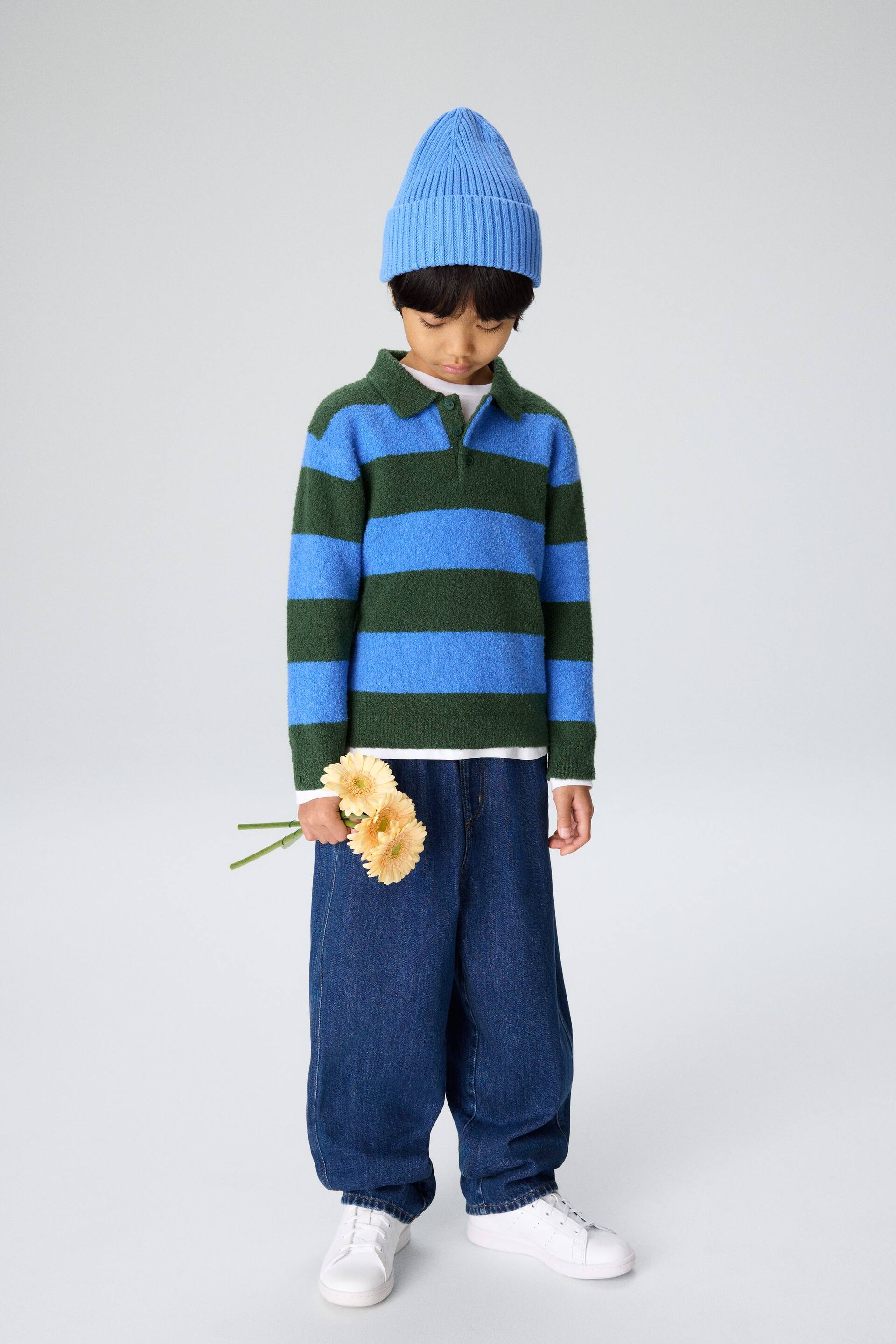 Zobraziť väčší obrázok: Oblečenie je prezentované spredu, obsahuje rebrovanú modrú čiapku a chlpatý pruhovaný polo sveter s modrými a tmavozelenými horizontálnymi pruhmi a dvojgombíkovým zapínaním, vrstvený cez biele tričko s dlhým rukávom. Tmavomodré široké džínsové nohavice sú nosené s bielymi šnurovacími teniskami.