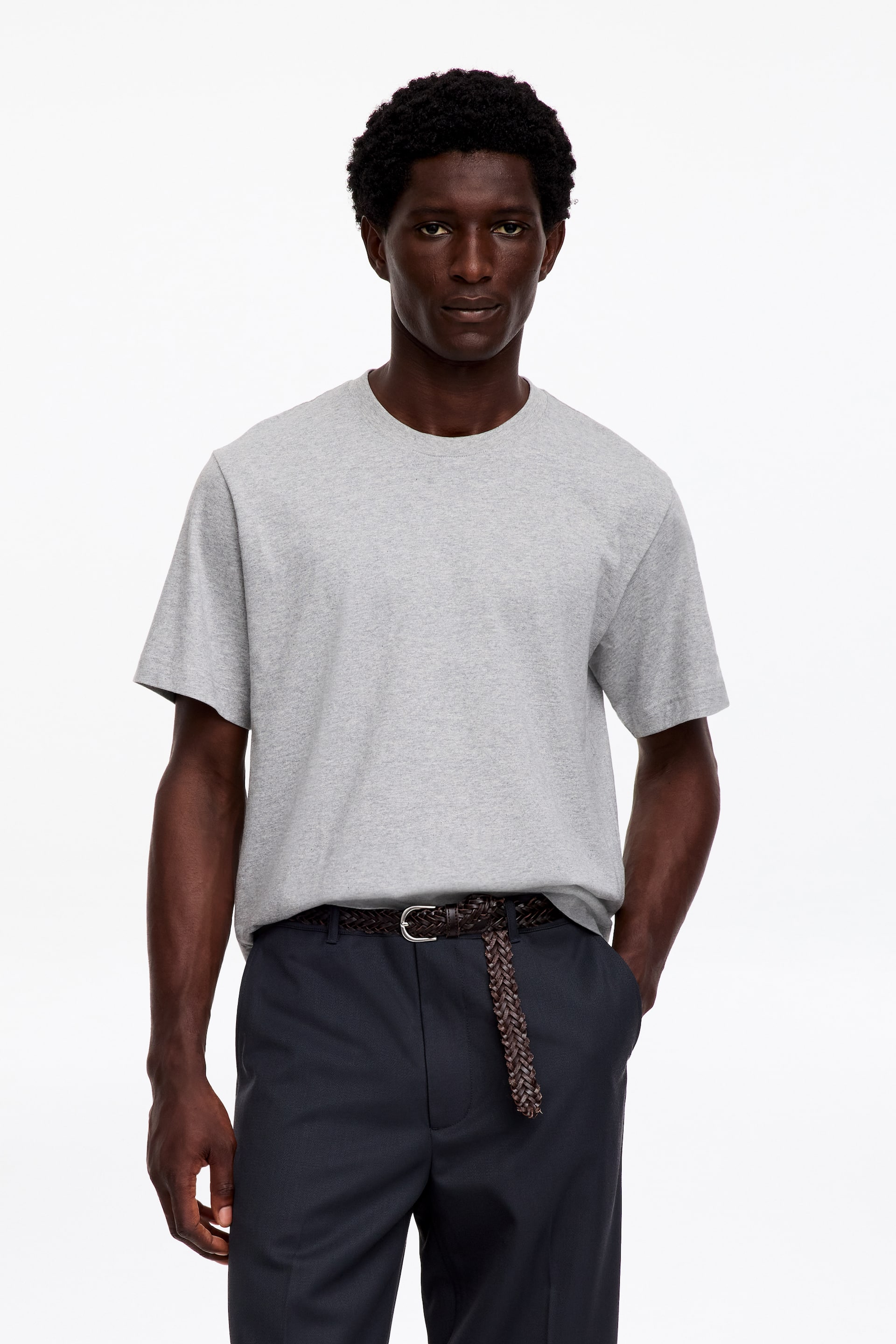 Visualizza l'immagine ingrandita: La modella indossa una T-shirt grigio chiaro con scollo a girocollo, collo a coste, vestibilità rilassata e maniche di lunghezza standard. La T-shirt è infilata in pantaloni scuri, con una cintura intrecciata marrone visibile in vita.