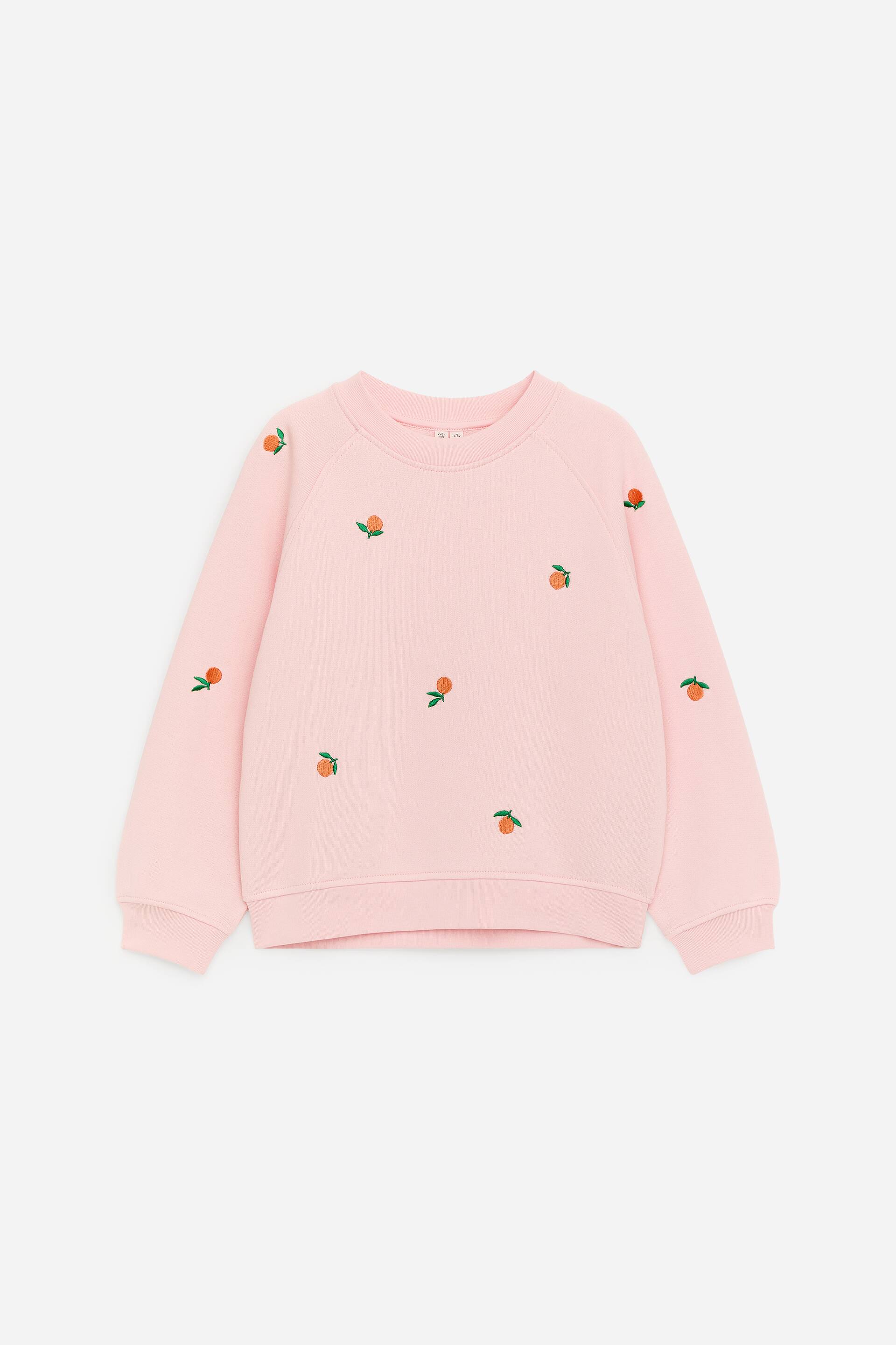 Visa större bild: En rosa sweatshirt med rund hals, långa raglanärmar, ribbade muddar och en ribbad fåll, pryds med utspridda broderade orange motiv, var och en med ett grönt blad, över framsidan och ärmarna.