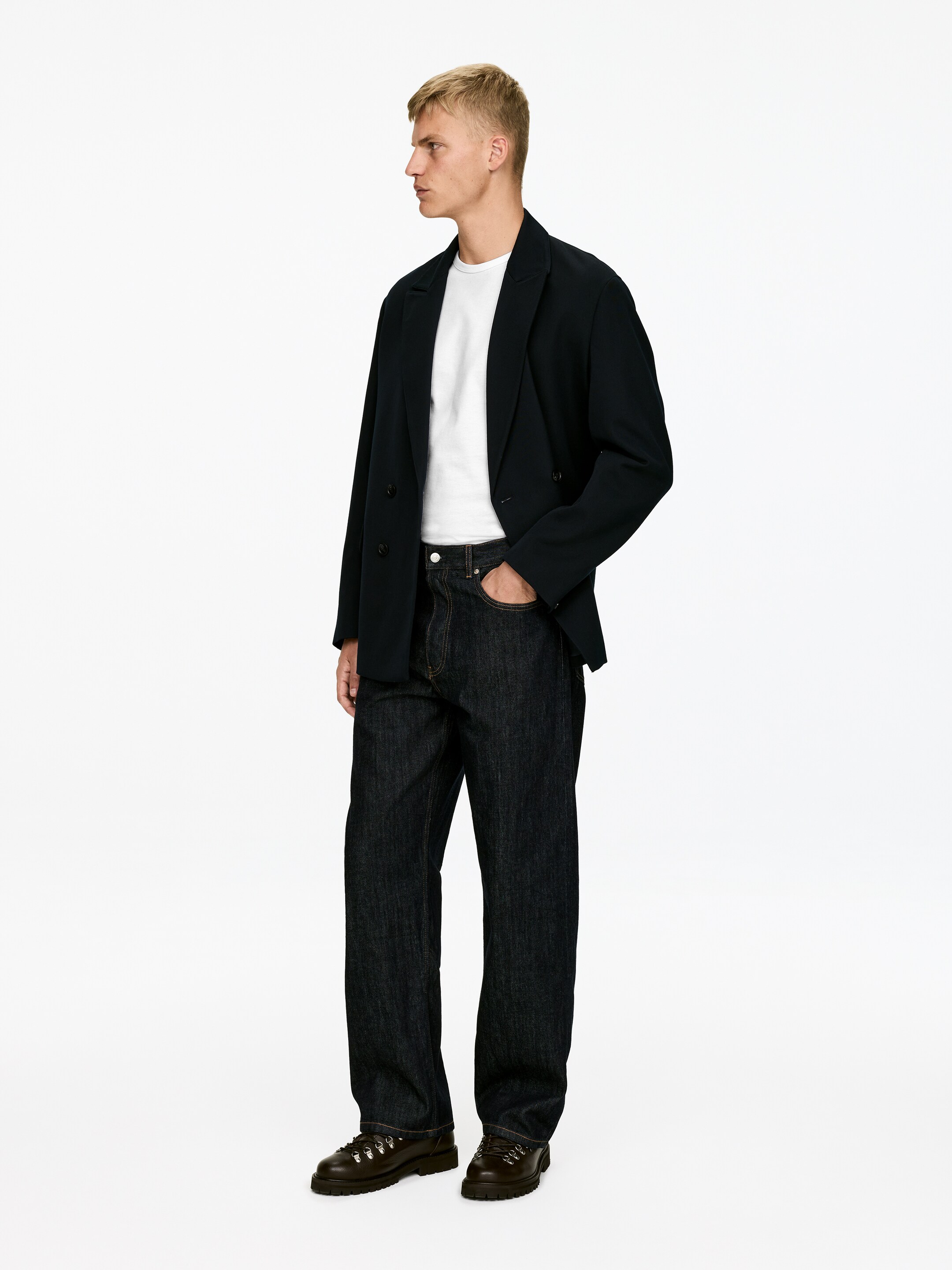 Agrandir l'image: Une veste bleu foncé à double boutonnage avec des revers en pointe est portée déboutonnée sur un t-shirt à col ras du cou blanc, associée à un jean à jambes larges et à délavage foncé et des chaussures à lacets marron foncé avec des semelles crantées. L'ensemble est présenté de profil, tourné vers la droite.