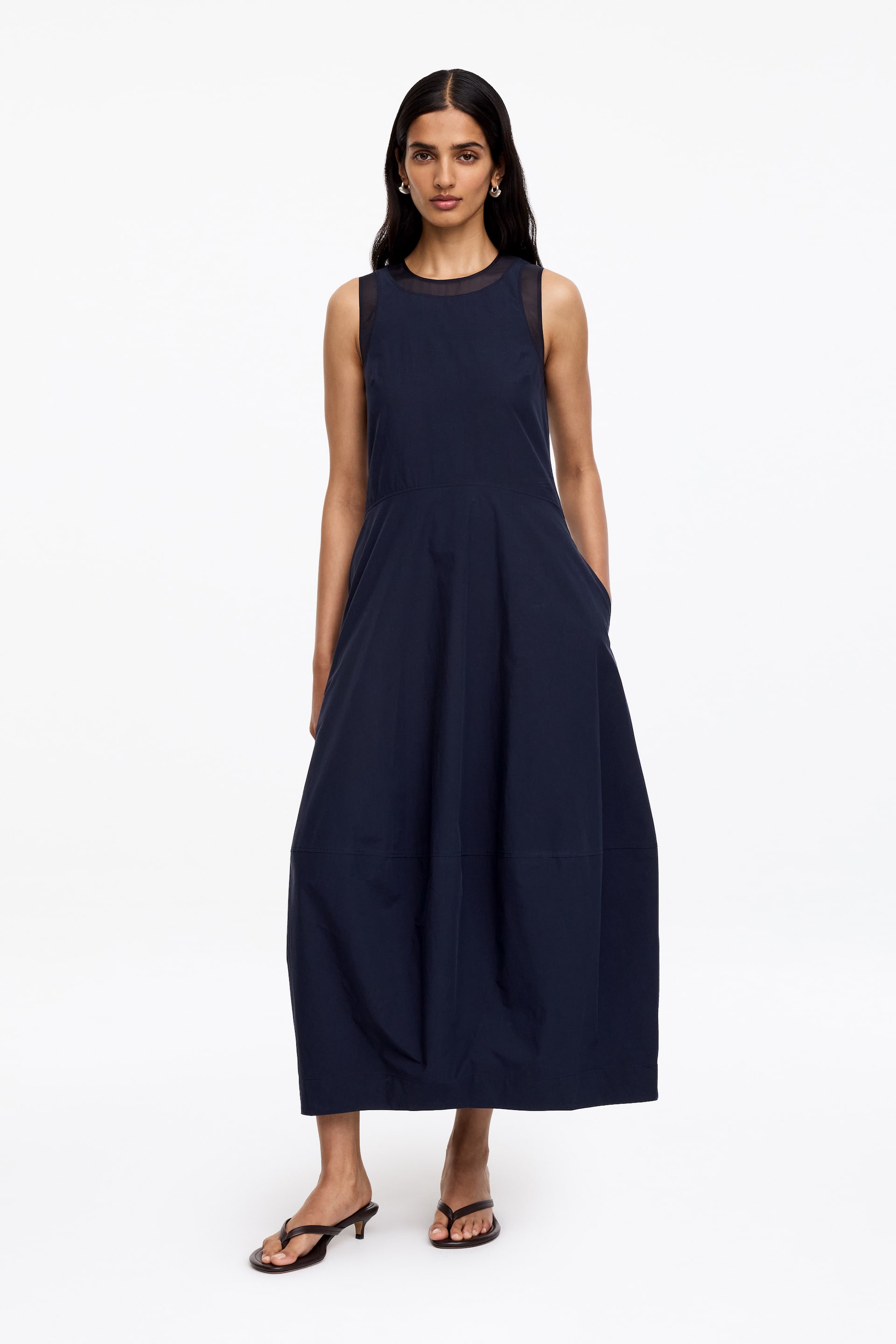 Grösseres Bild ansehen: Ein ärmelloses Midikleid in Marineblau mit einem transparenten, runden Joch am Ausschnitt. Das Kleid ist mit einem figurbetonten Oberteil gestaltet und verfügt über einen vollen, ausgestellten Rock, der in Midilänge fließt und sichtbare Paneele sowie seitliche Taschen enthält.