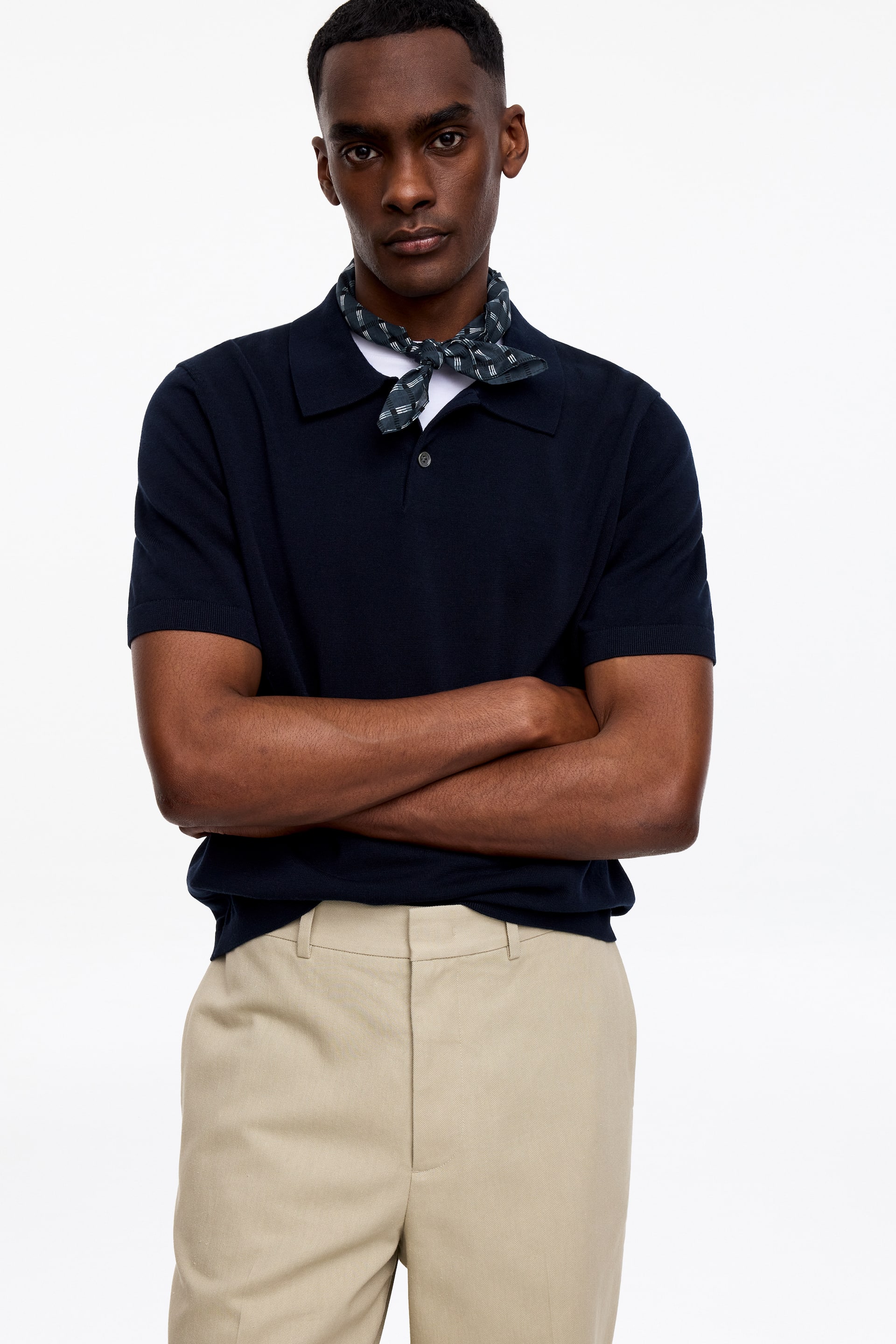 Grösseres Bild ansehen: Ein marineblaues, kurzärmeliges Strick-Poloshirt wird von vorne präsentiert, mit einem Kragen, einer Zwei-Knopf-Leiste und gerippten Bündchen. Es ist über einem weißen Unterhemd getragen, mit einem dunkel gemusterten Halstuch um den Hals gebunden und mit beigefarbenen Flat-Front-Hosen kombiniert.