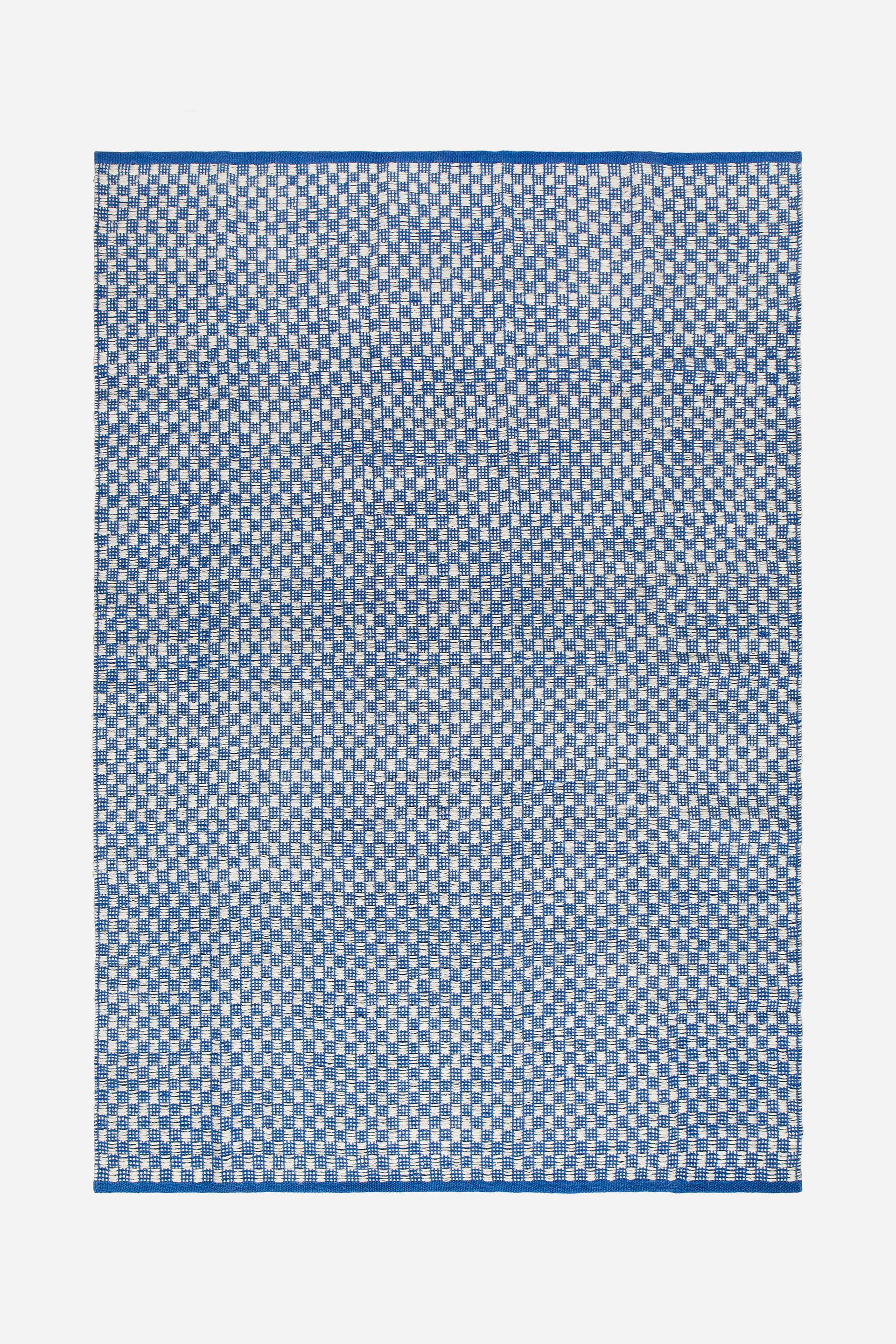 Se større billede: Et rektangulært fladvævet tæppe viser et mønster af små, vekslende blå og hvide firkanter. Dets øverste og nederste kanter er indrammet af et solidt blåt bånd.