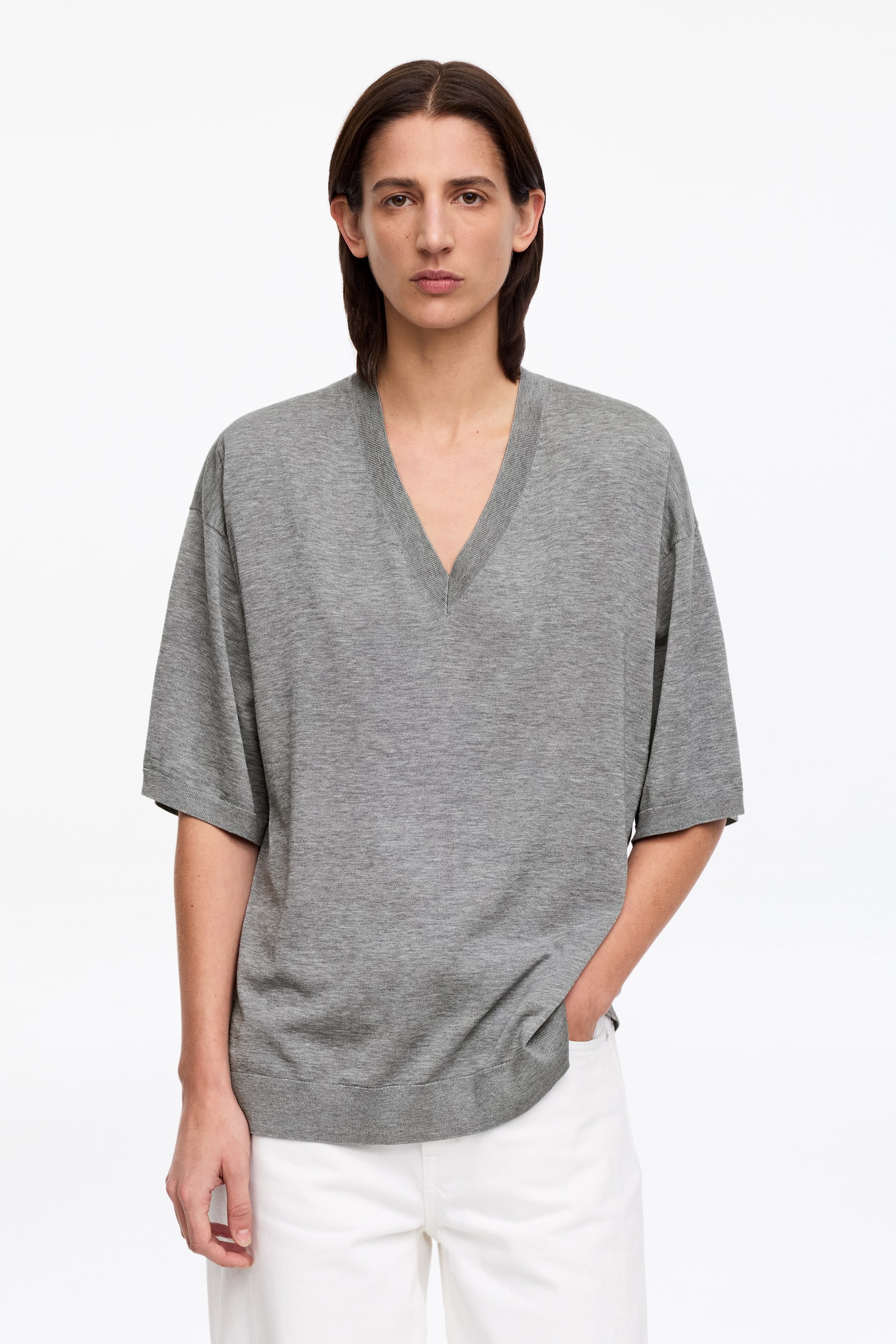 Ingrandisci l'immagine: La modella indossa un top grigio a maniche corte in maglia. È tagliato con uno scollo a V e cade in una vestibilità ampia. La modella sta mettendo una mano nella tasca dei pantaloni bianchi, che sono abbinati al top.