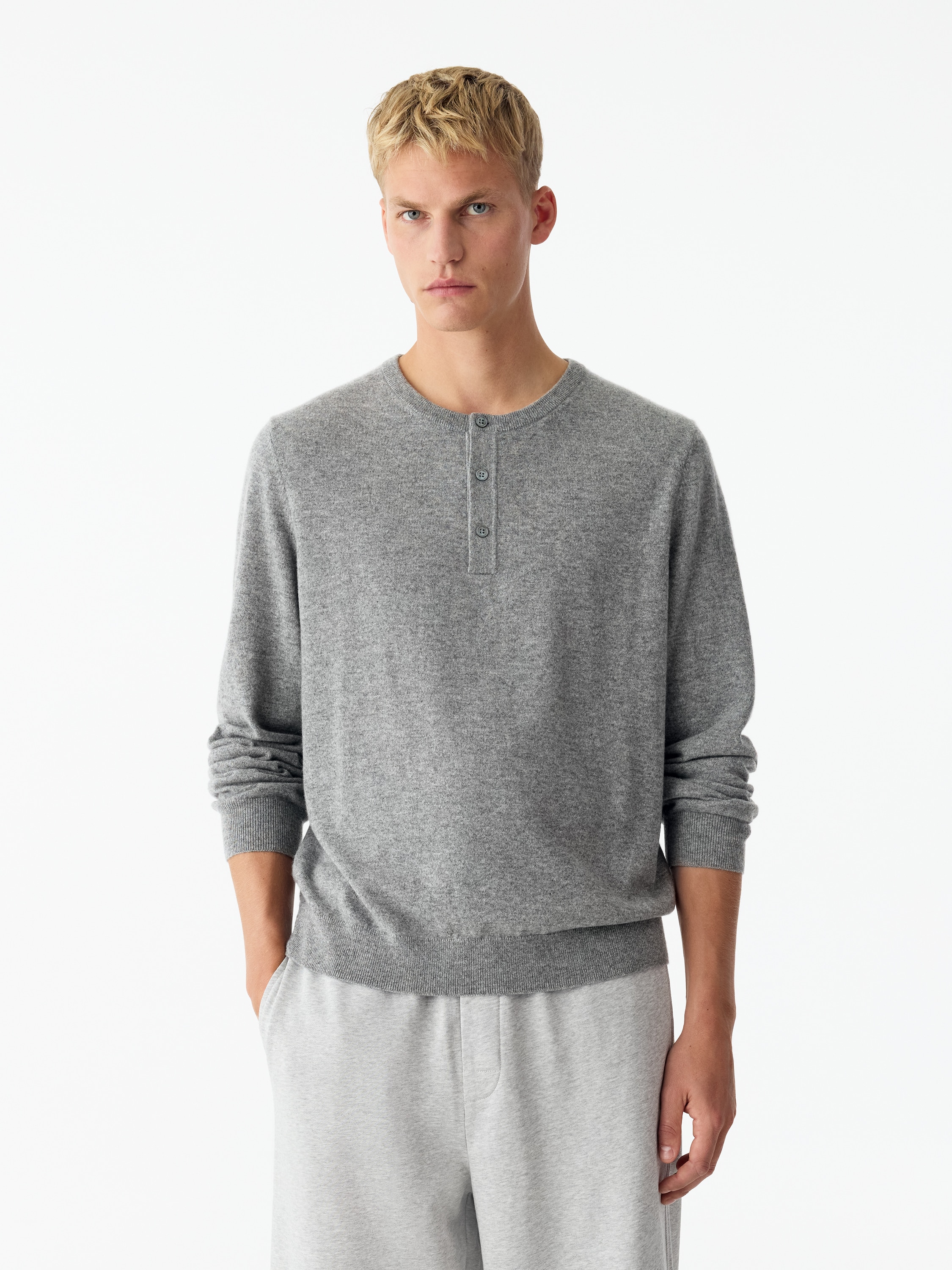 Un maglione henley mélange grigio chiaro è mostrato frontalmente, con scollo a costine a doppio strato e abbottonatura a tre bottoni con bottoni chiari. Il capo a maniche lunghe presenta una texture a maglia fine, rifinita con polsini a costine e fondo a costine.