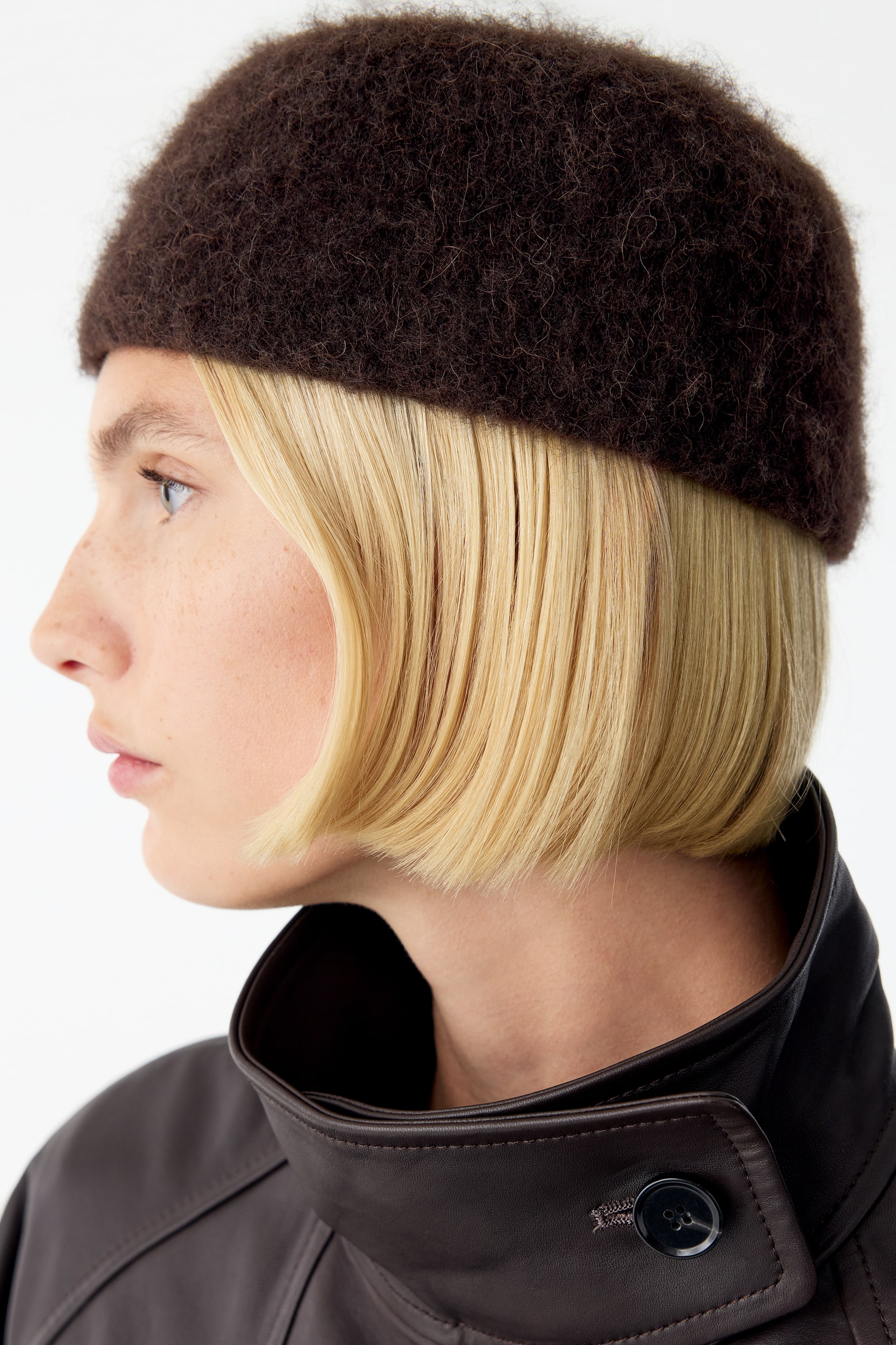 View larger image: A dark brown, textured knitted hat with a rounded pillbox shape is being worn by the model in profile. The fuzzy-textured hat is styled with a dark brown leather jacket featuring a high, buttoned collar.