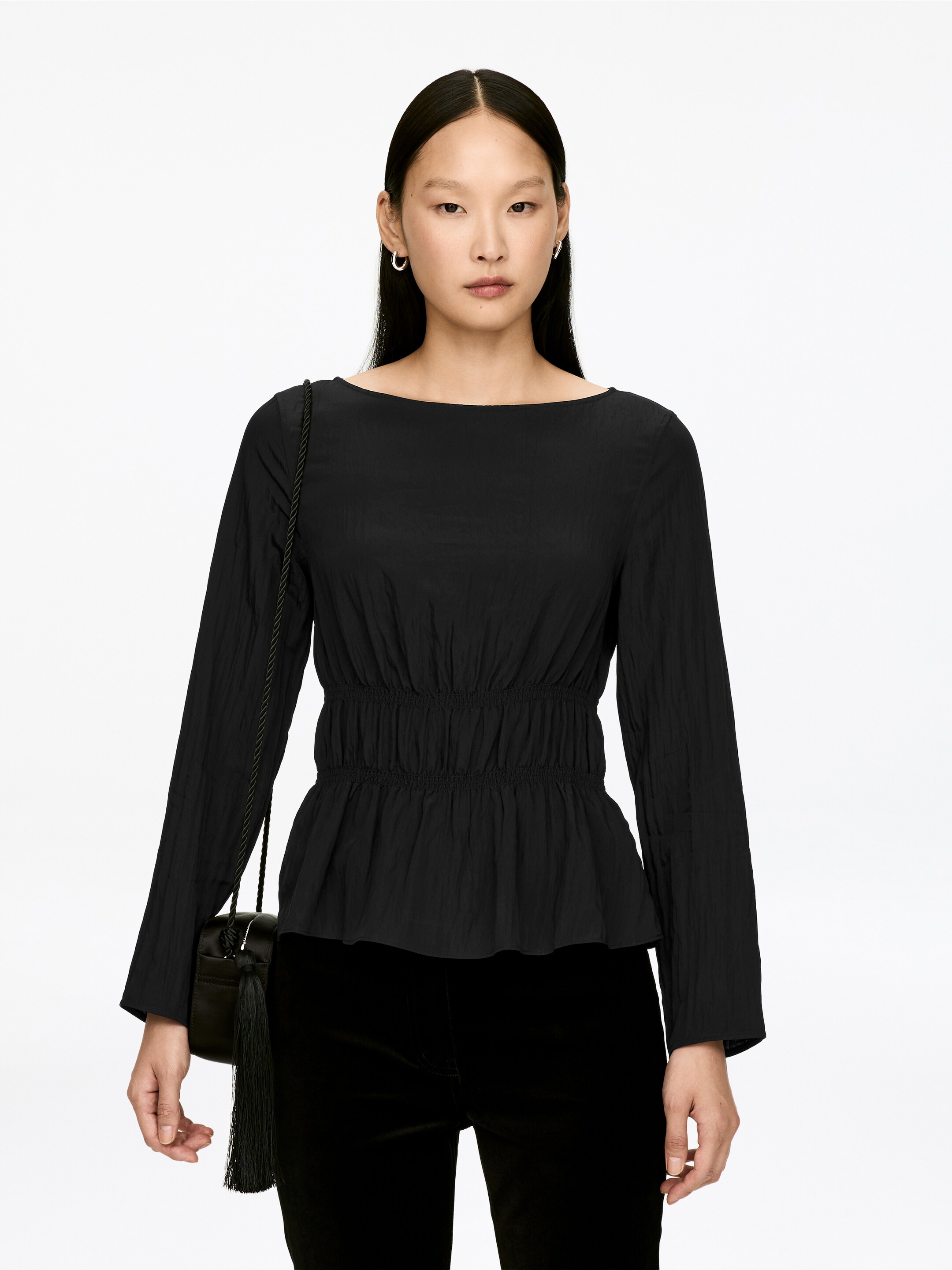 View larger image: The black top is featuring a boat neckline and long sleeves. It is designed with a shirred waistline that is flowing into a peplum hem, presenting a subtle crinkled texture throughout.