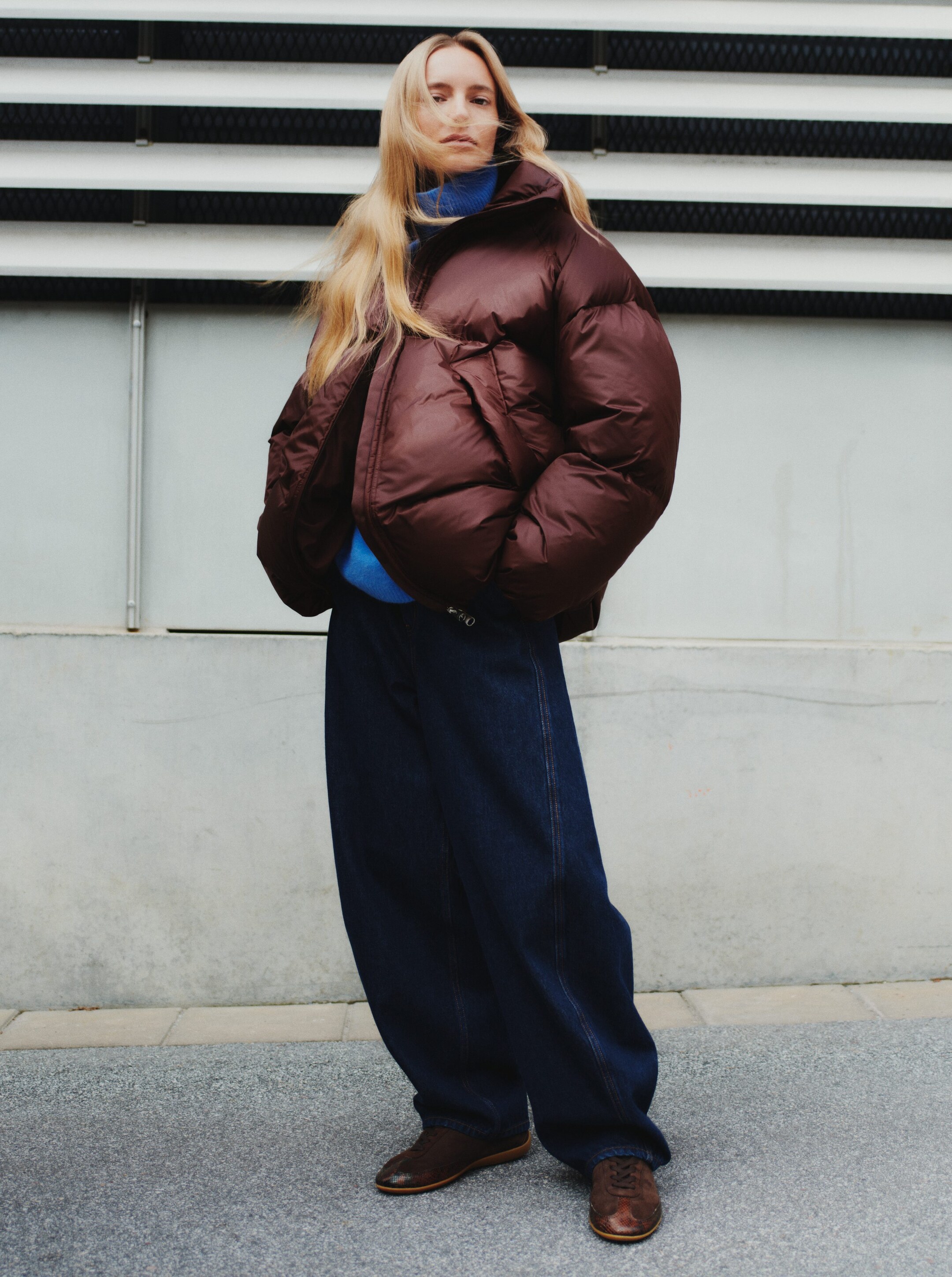 Größeres Bild anzeigen: Eine voluminöse, kurz geschnittene braune Steppjacke mit einem hohen Stehkragen und Seitentaschen wird offen über einem blauen Rollkragenpullover getragen.