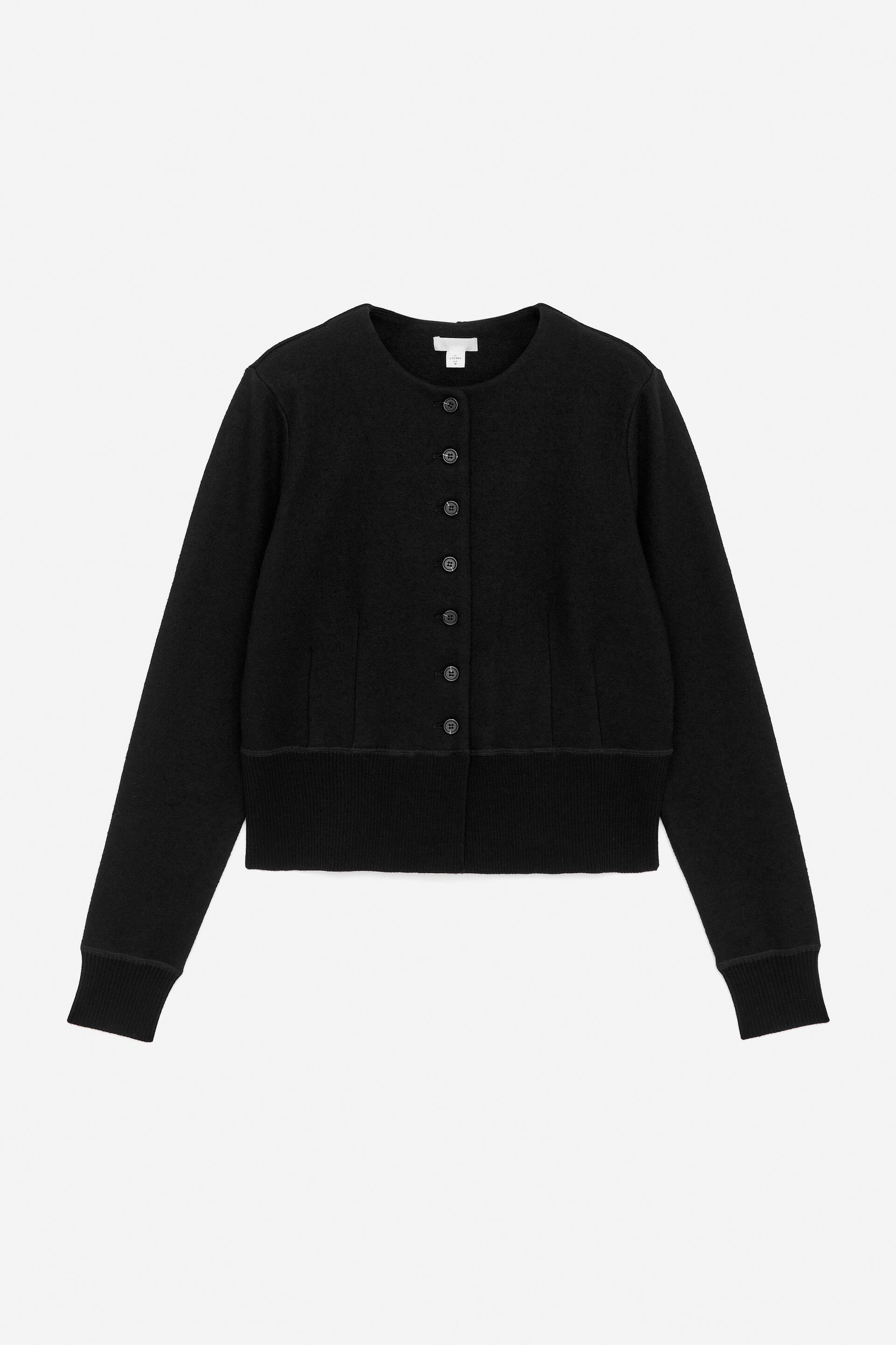 Vis større bilde: En svart cardigan presenteres fra forsiden, med en rund halslinning og en full knappestolpe med syv mørke, sirkulære knapper. Den er designet med lange ermer, ribbestrikkede mansjetter og en ribbestrikket kant nederst, med en horisontal søm plassert over kanten.