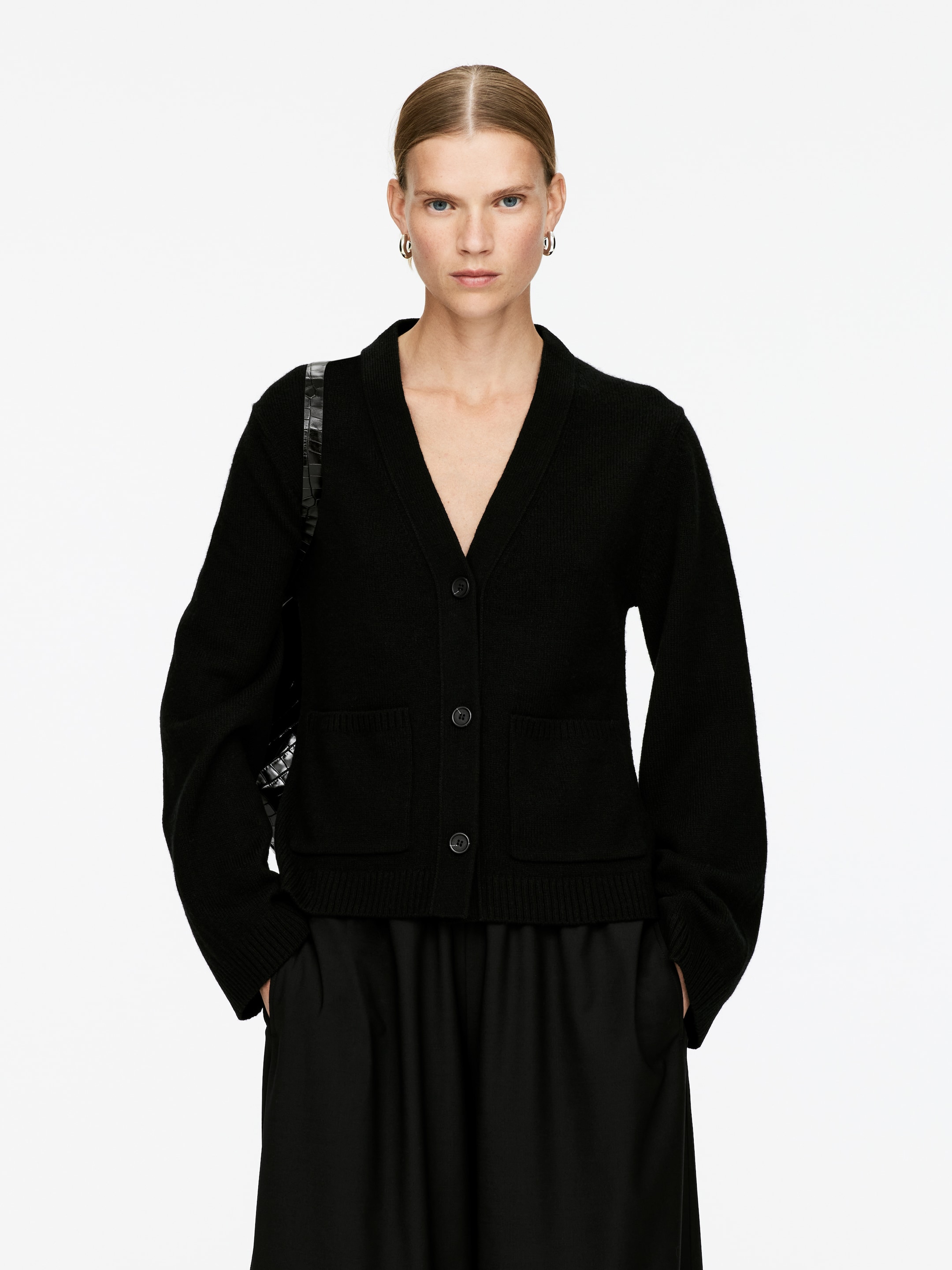View larger image: A black cardigan is being presented straight on, featuring a V-neckline with a shawl collar. It has a three-button front closure, two patch pockets, and is finished with ribbed cuffs and a ribbed hem, being styled over black wide-leg pants.