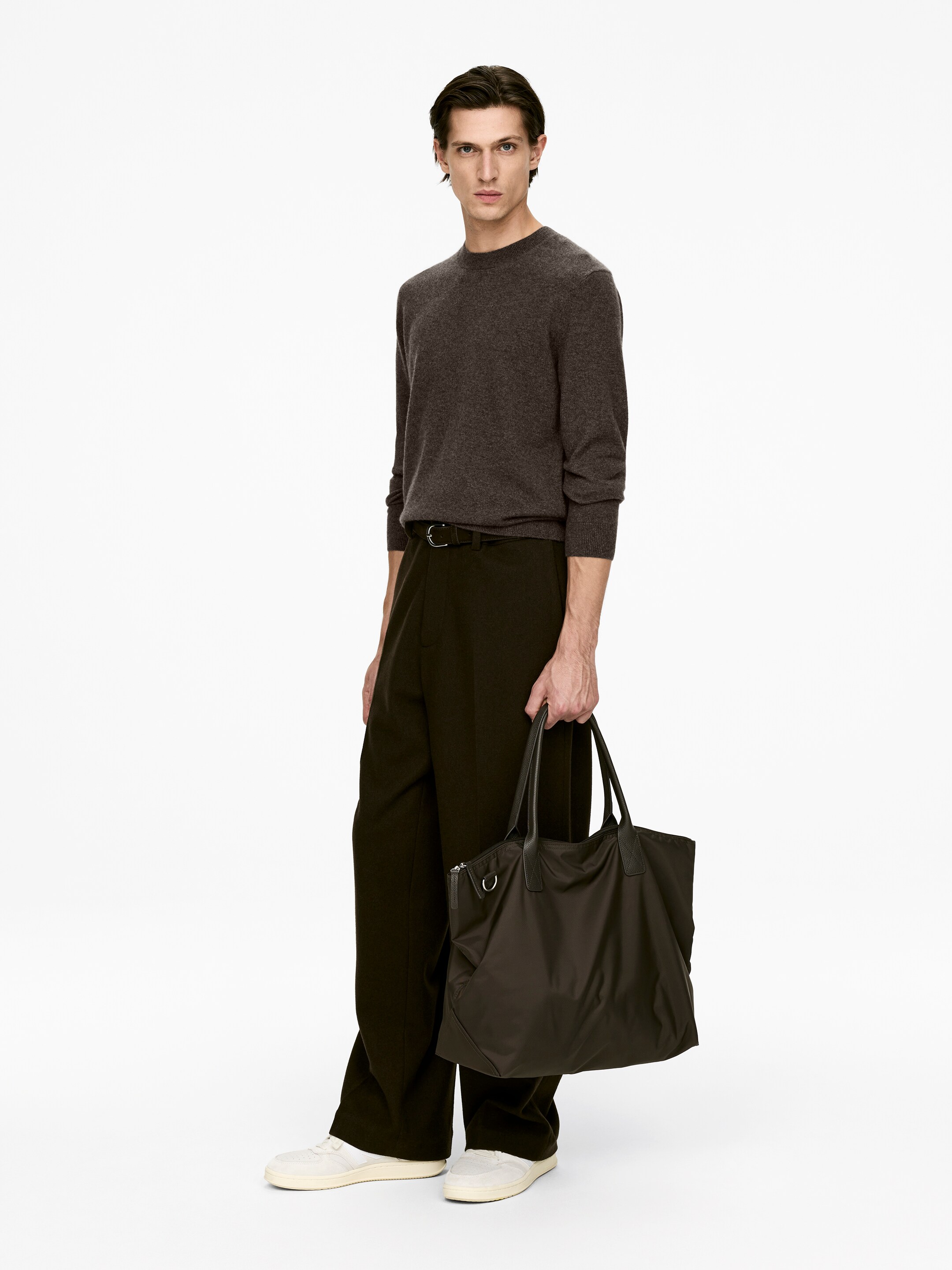 View larger image: A dark brown tote bag is being held, facing slightly towards the viewer. It is featuring a soft, flexible fabric construction with two darker top handles and a visible zipper closure along the top edge.