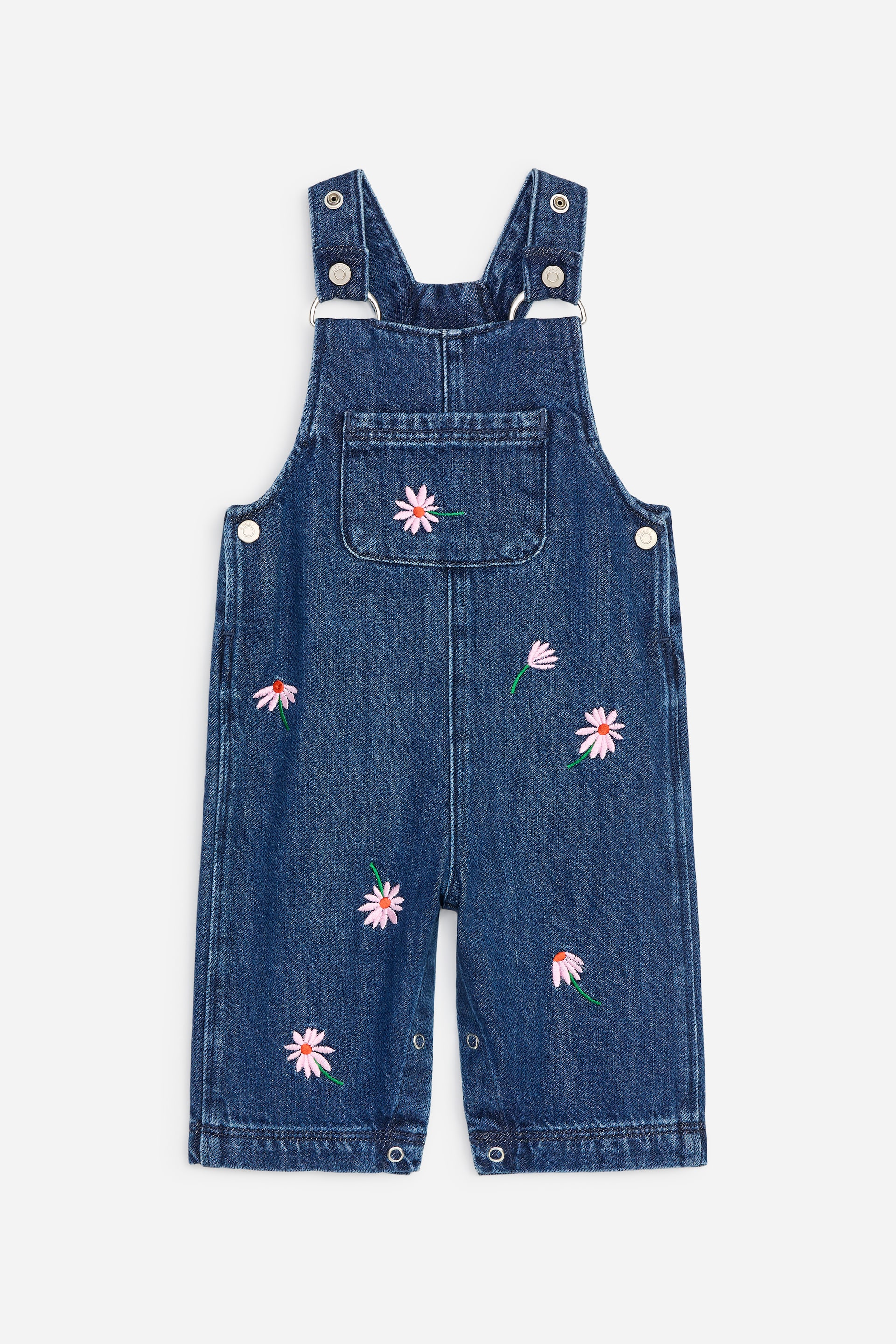 Agrandir l'image: Le pantalon salopette en denim est montré de face. Il présente des fleurs brodées rose et blanches de type marguerite, une poche poitrine, des bretelles réglables et des boutons-pression le long de l’intérieur de la jambe.