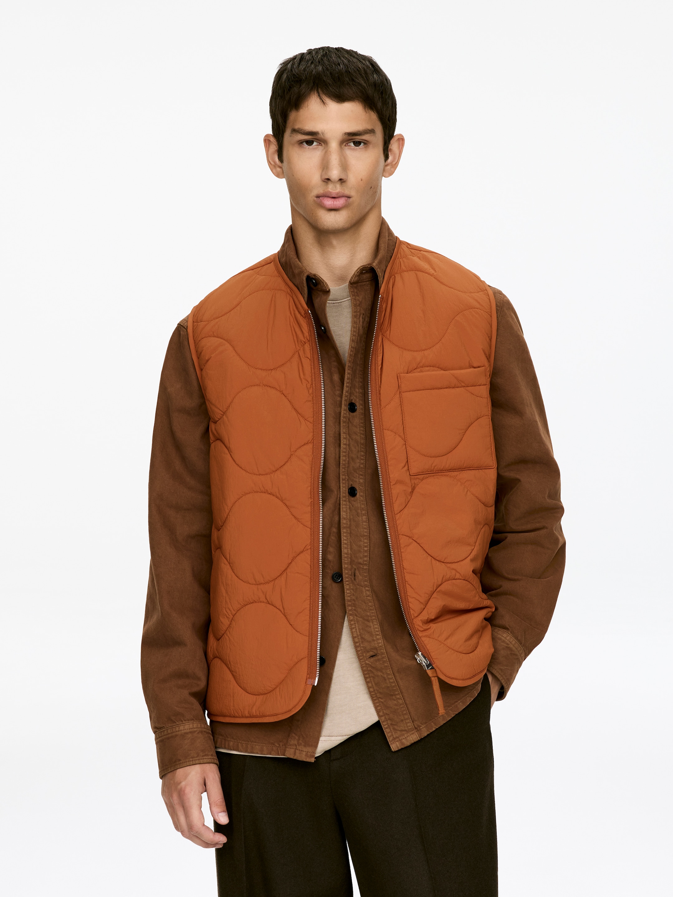 Agrandir l'image: Un gilet matelassé orange à l'avant présente un motif matelassé ondulé, un zip frontal argenté et une poche plaquée sur la poitrine gauche du porteur. Il est porté ouvert sur une chemise à col marron, révélant son ourlet courbé.