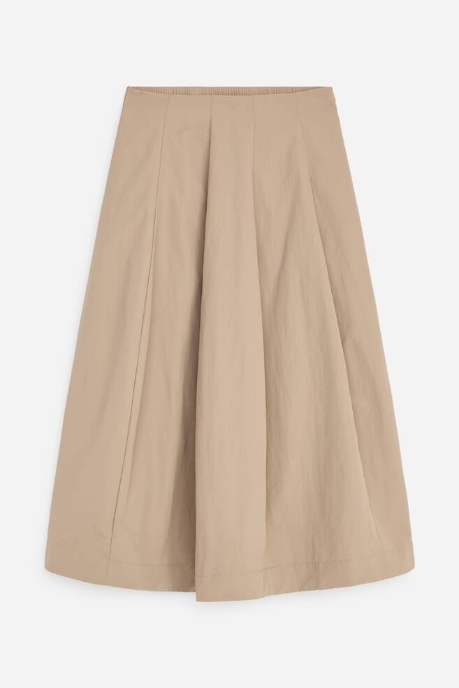 A beige A-line midi skirt is hanging from a front angle, showcasing a panelled construction with deep vertical pleats that create a flared, voluminous silhouette. The skirt features a flat front waistband with an elasticated back and a wide, topstitched hem. - 1