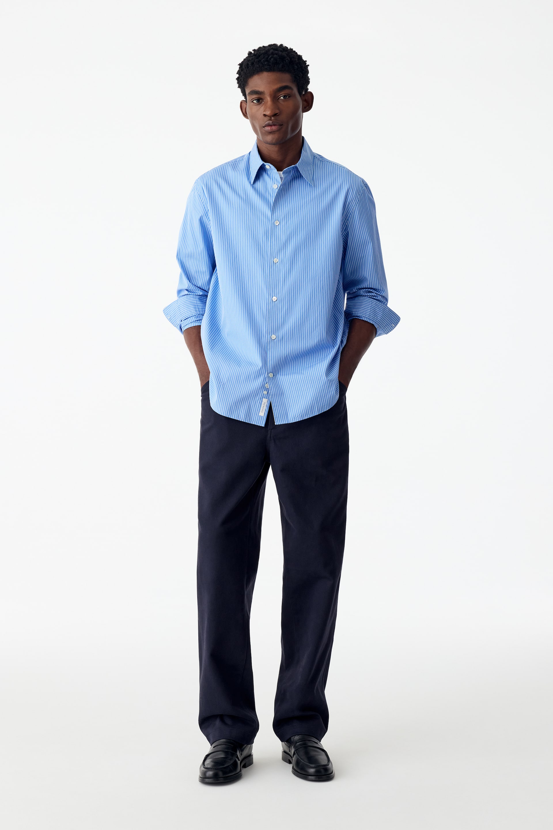 Grotere afbeelding bekijken: Een model draagt een relaxte broek van katoen en lyocell in donker marineblauw, terwijl het rechtop staat met de handen in de zakken. De broek met rechte pijpen en losse pasvorm is gestyled met een blauw-wit gestreept hemd met knopen en zwarte leren loafers.