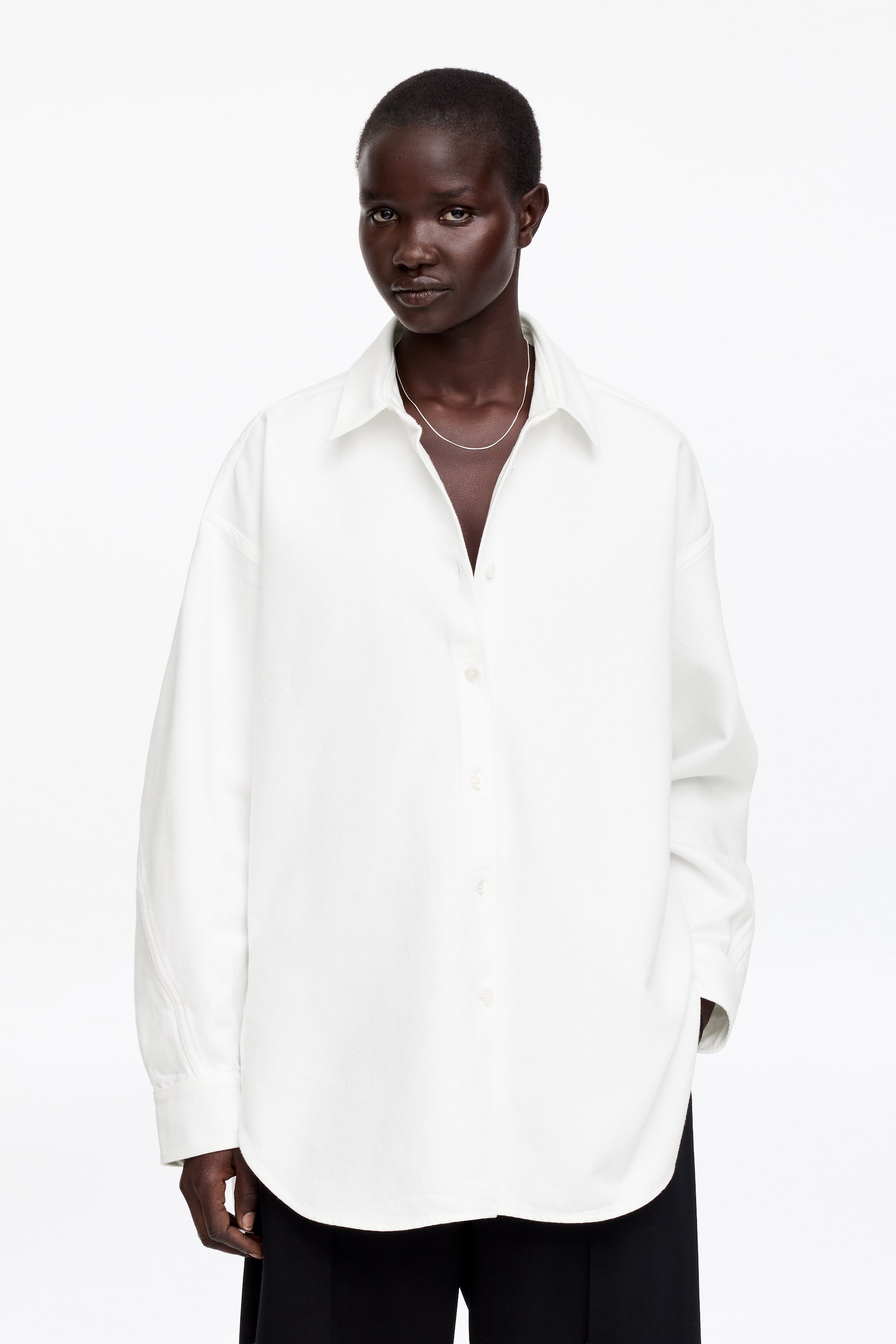 Grösseres Bild ansehen: Ein weißes, übergroßes Hemd mit Knopfleiste wird präsentiert, nach vorne gerichtet. Es verfügt über einen Kentkragen mit offenem Ausschnitt, eine sichtbare Knopfleiste entlang der Vorderseite und lange Ärmel mit geknöpften Manschetten. Das Hemd zeigt einen abgerundeten Saum und wird ungetuckt über dunklen Hosen gestylt.
