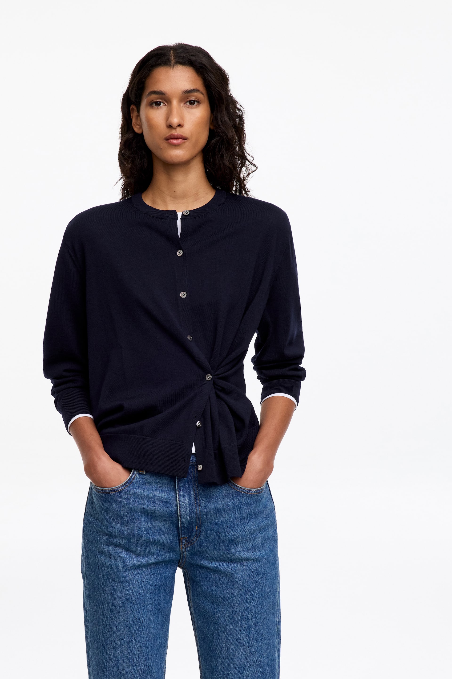 Größeres Bild ansehen: Eine marineblaue Strickjacke mit Rundhalsausschnitt, Knopfleiste vorne und asymmetrischem Twist-Detail an der Taille wird vom Model getragen. Die Strickjacke ist über einem weißen Oberteil getragen und mit blauen Jeans kombiniert.