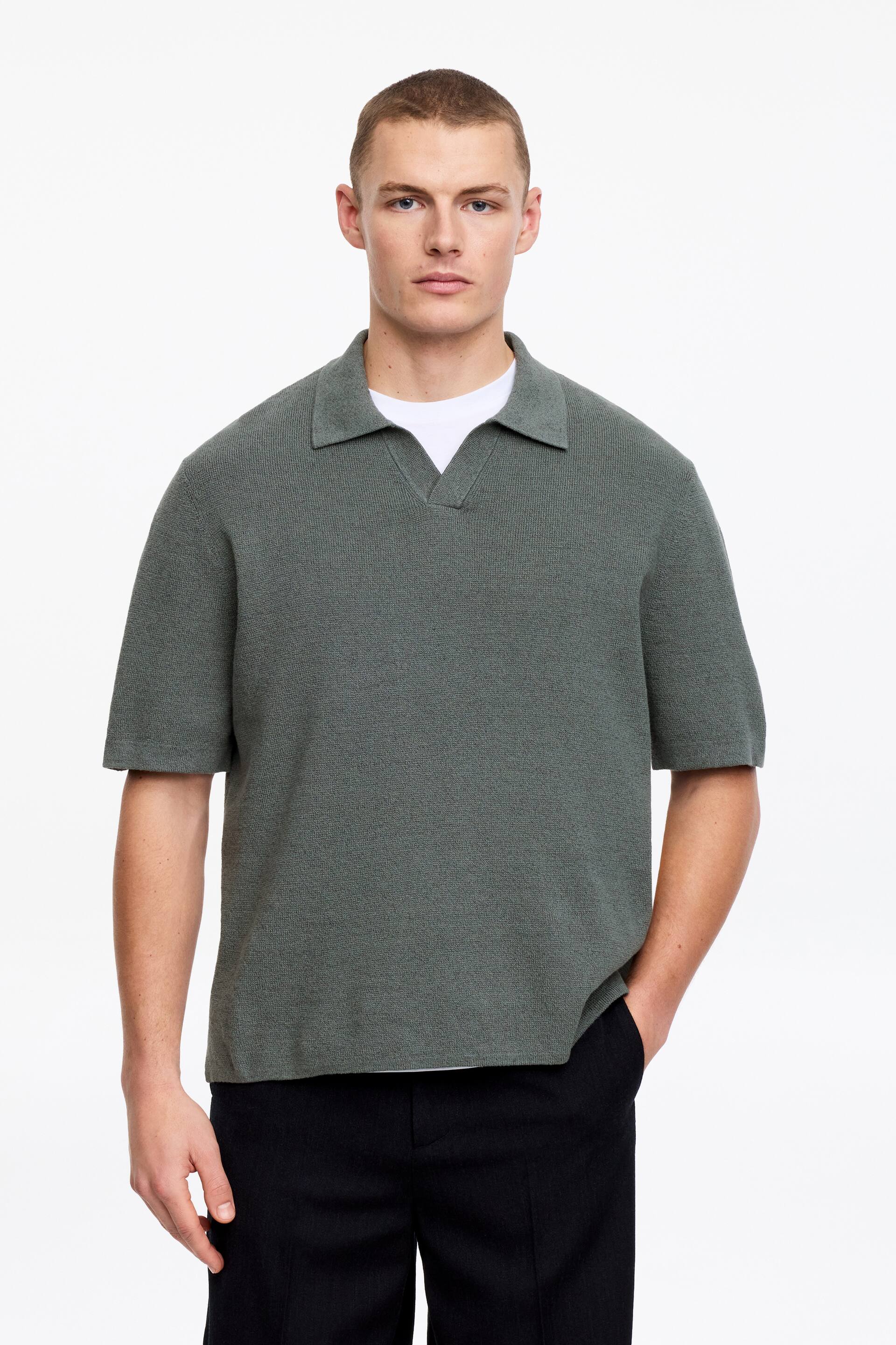 Προβολή μεγαλύτερης εικόνας: Ένα σκούρο πράσινο πλεκτό πόλο πουκάμισο με υφή διαθέτει γιακά με άνοιγμα, V λαιμόκοψη και κοντά μανίκια. Φοριέται πάνω από ένα λευκό μπλουζάκι, ορατό στη λαιμόκοψη και στο στρίφωμα.