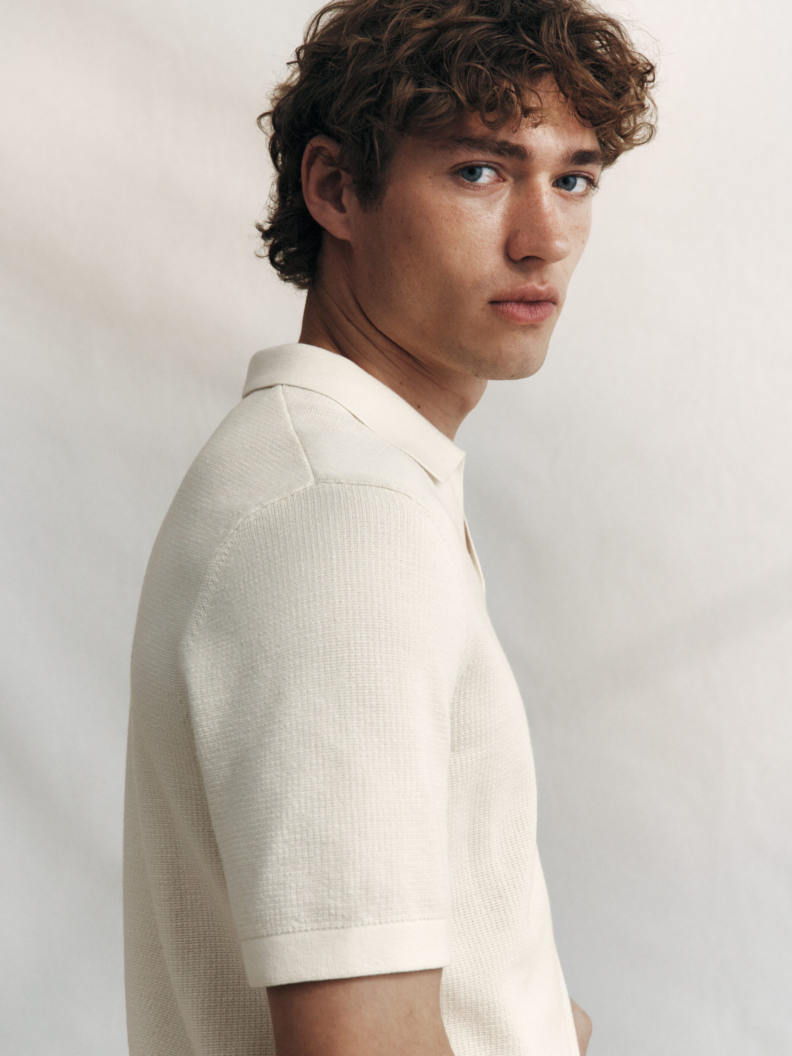 A light cream polo shirt is showcasing its front view, featuring a textured knit surface. It is designed with short sleeves and a V-neck spread collar, maintaining a regular fit.