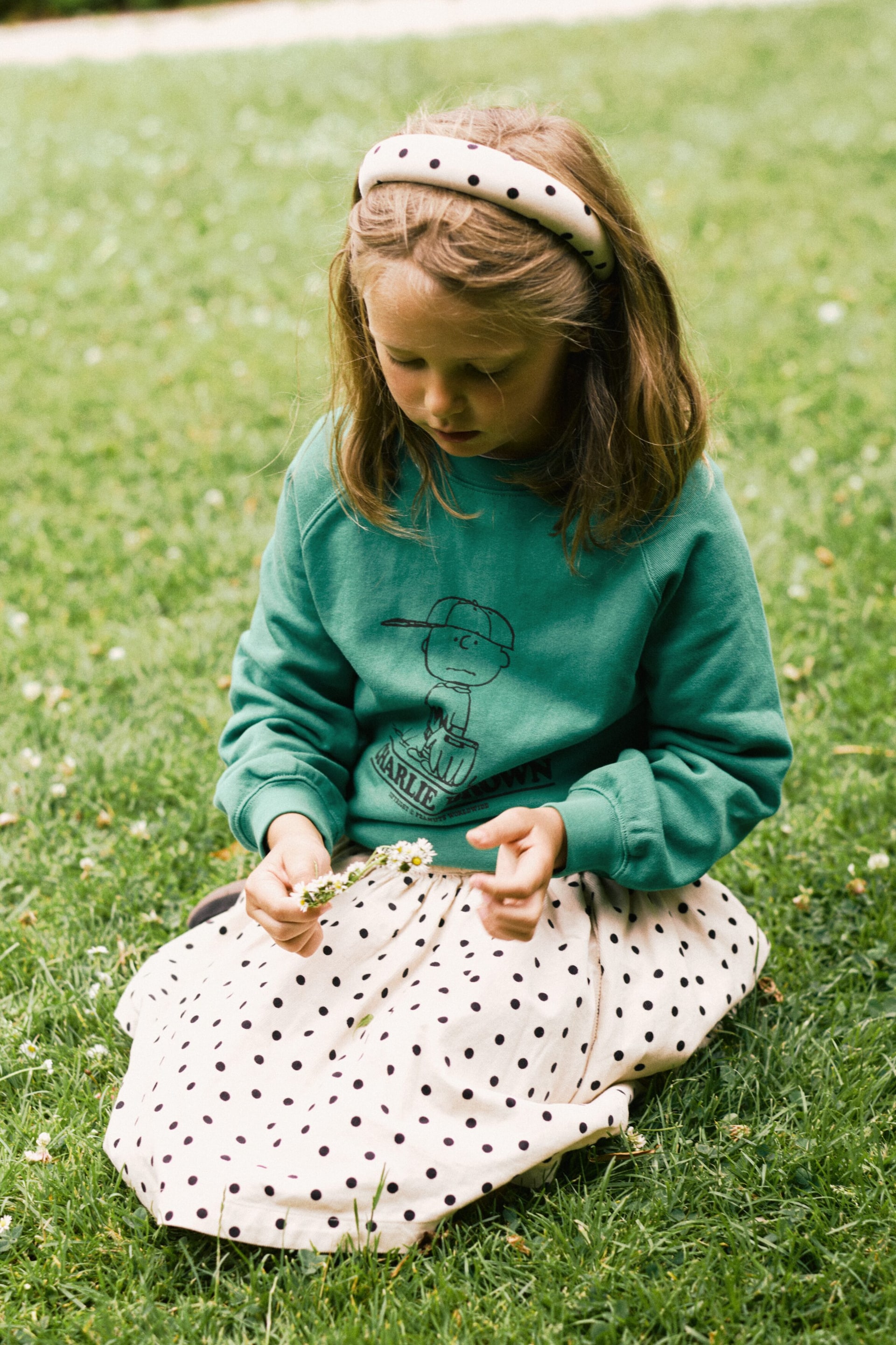 Visa större bild: En grön sweatshirt med rund hals visas framifrån och har en svart linjeteckning av en figur med keps samt texten ”CHARLIE BROWN”, tillsammans med synliga ribbade muddar och fåll. En hel beige kjol med svarta prickar visas också och faller i rynkningar runt bärarens ben.