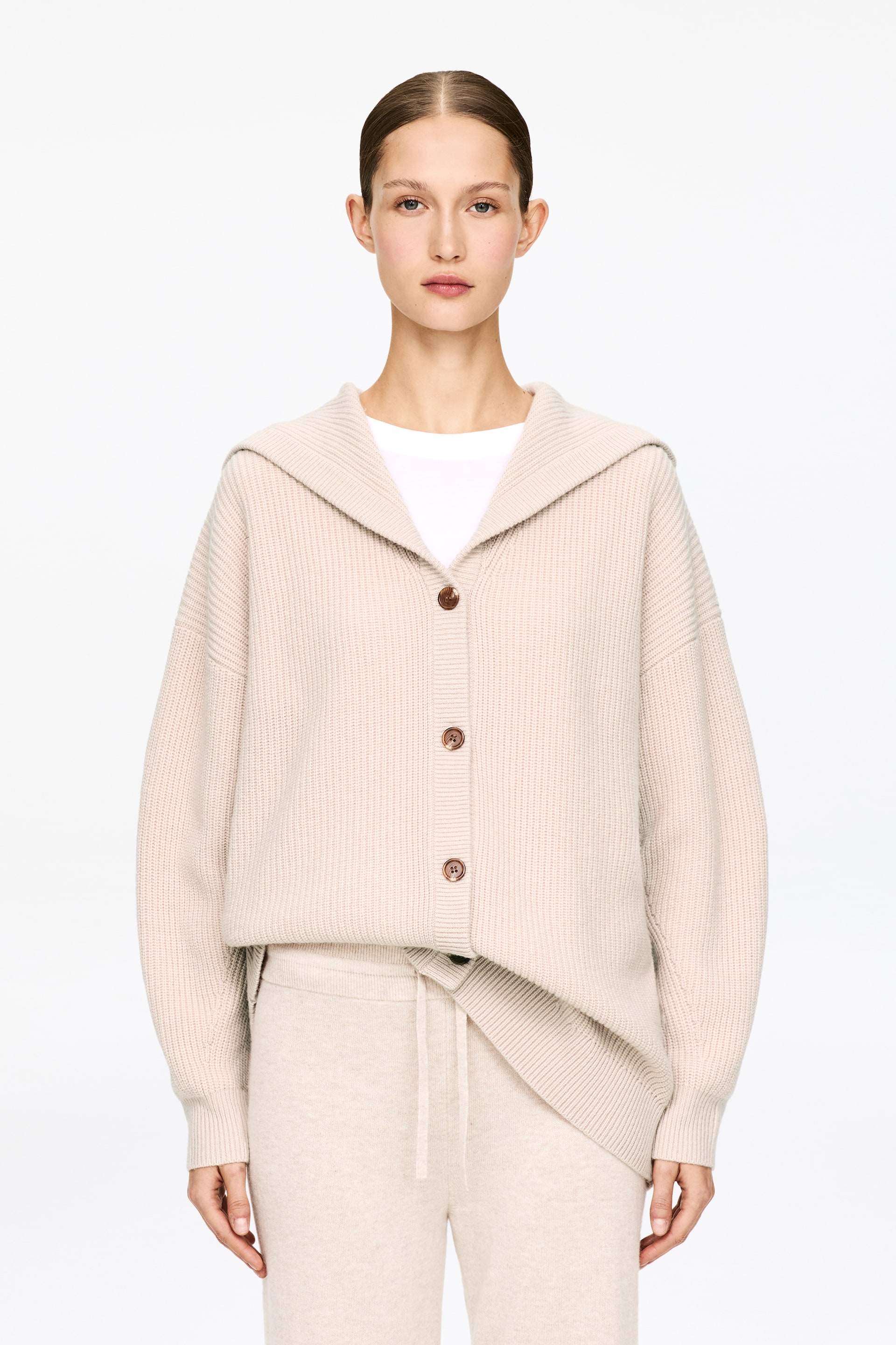 Visa större bild: En ljusbeige huvtröja med ballongärmar och ribbade kanter bärs av modellen. Koftan, med en avslappnad passform och bruna knappar, är uppknäppt över en vit t-shirt och matchas med ljusbeige byxor.