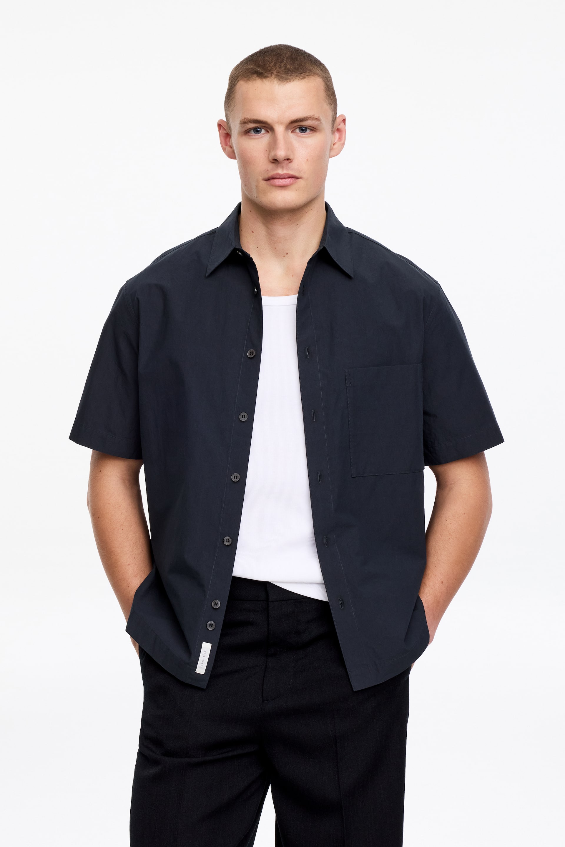 Grotere afbeelding bekijken: Het donkergekleurde overhemd met korte mouwen en kraag heeft een gespreide kraag, een enkele borstzak aan de linkerkant en donkere knopen langs de voorkant. Het wordt frontaal gepresenteerd, open gedragen over een wit onderhemd, en een klein wit label is zichtbaar aan de rechte onderzoom aan de linkerkant.